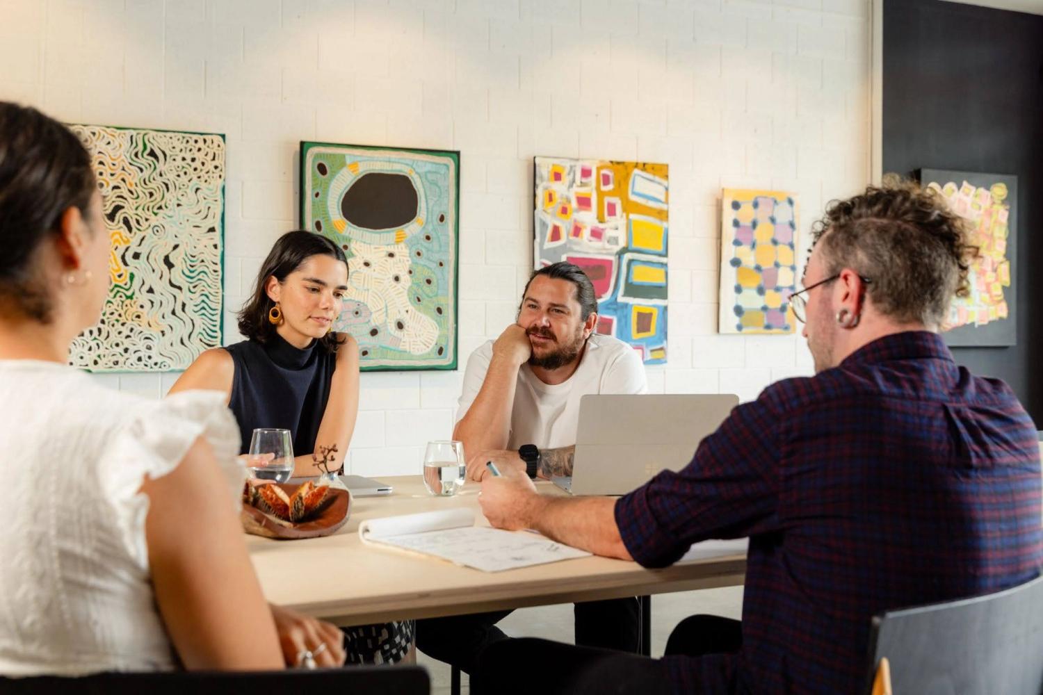 Four people meeting around a high bench with pens, paper and laptops, with art hanging in the background. 