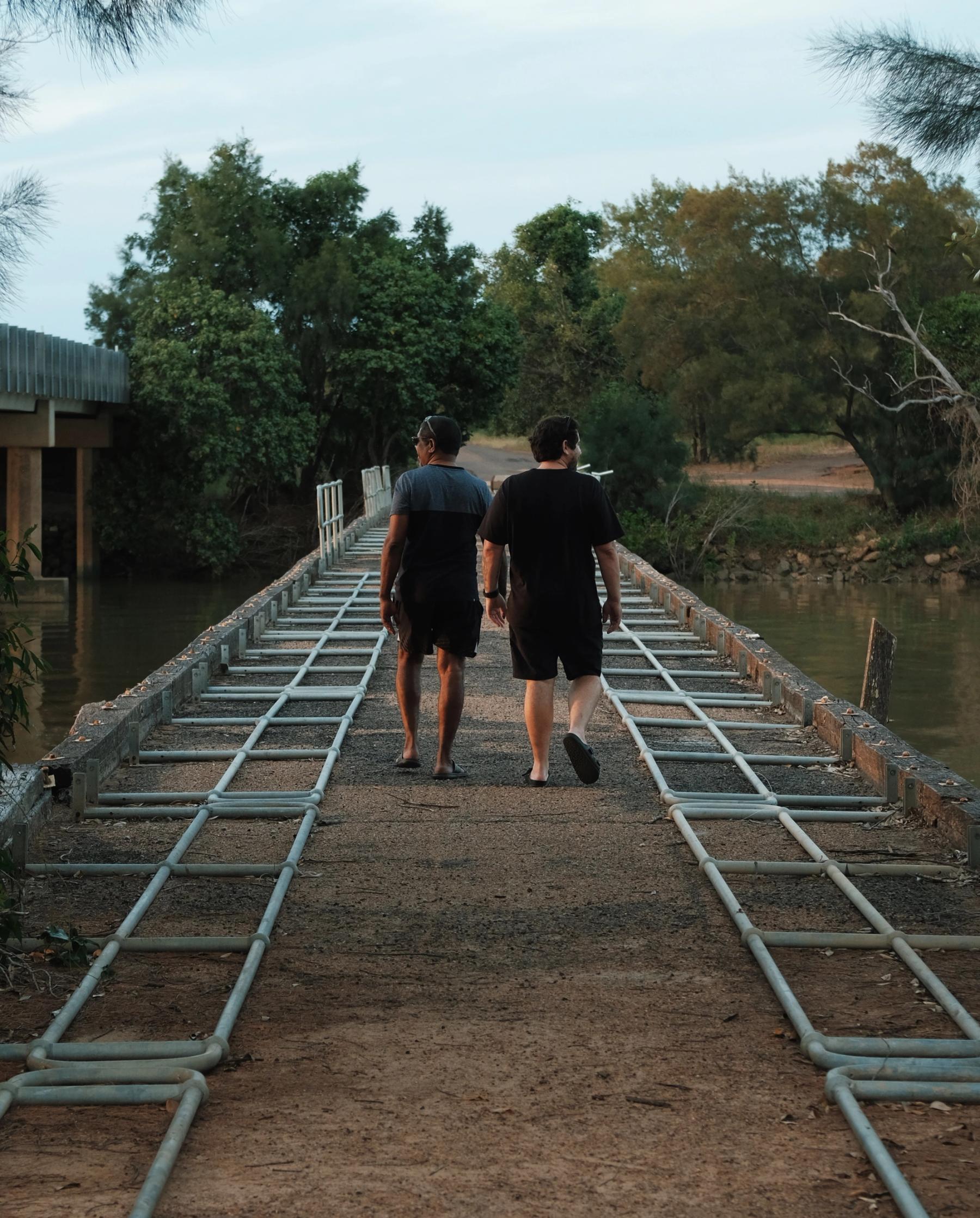 Two men walking away from the camera across a pedestrian bridge with a river below them and trees in front of them