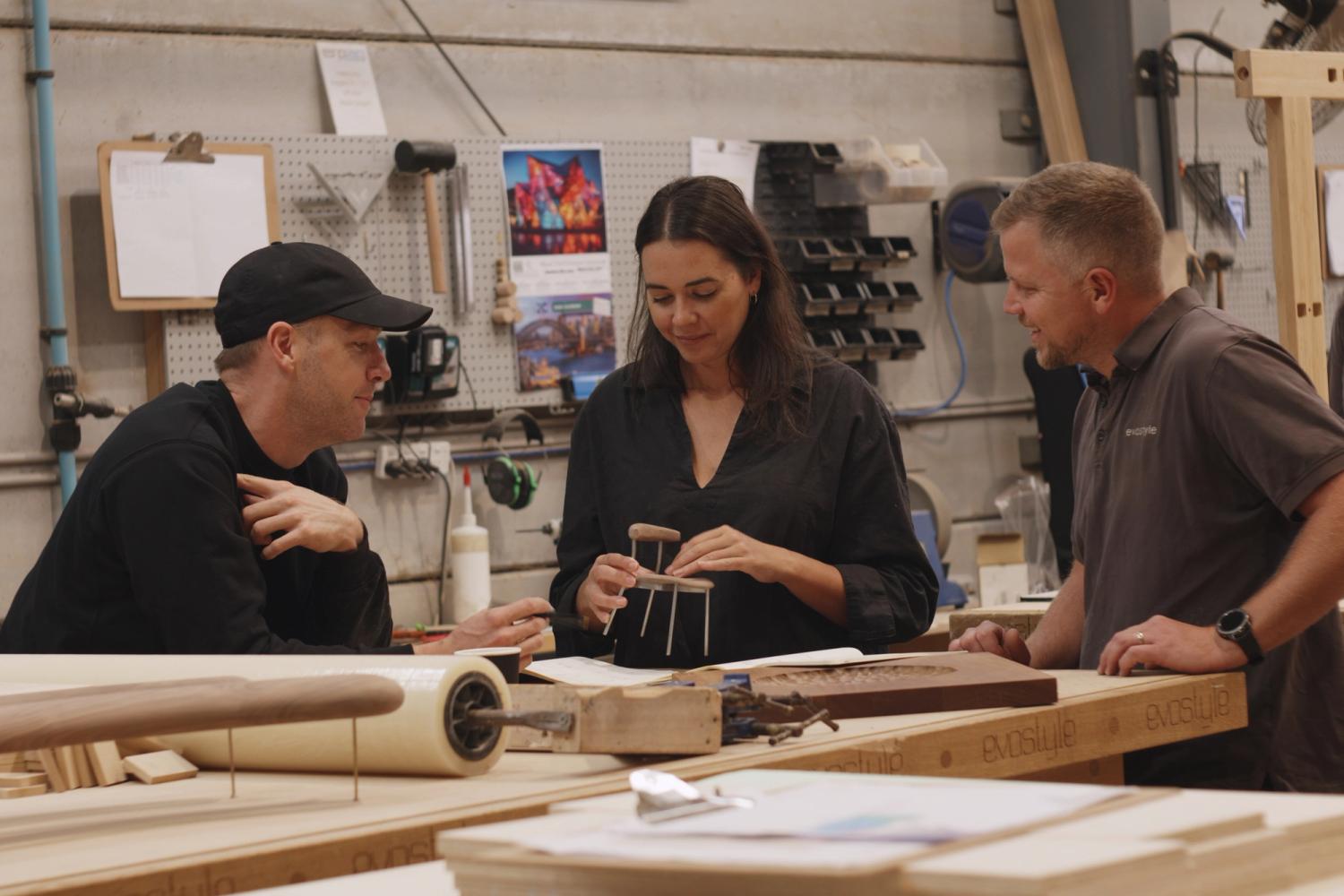 Three people around a timber worktable in a furniture workshop reviewing a stool design
