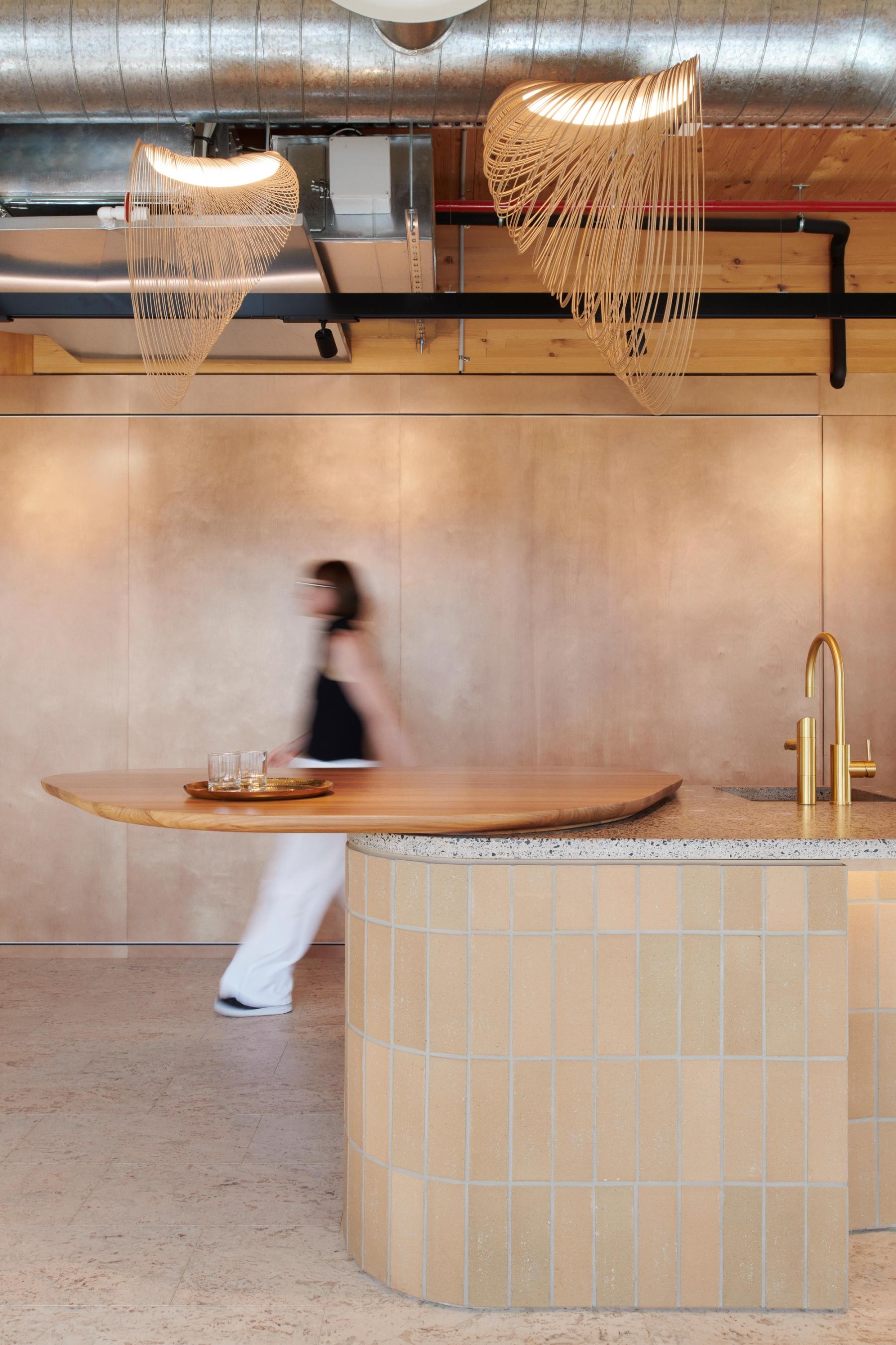 Office kitchen space with curved, tiled bench, gold fittings and wooden elements, a blurred person walks through.