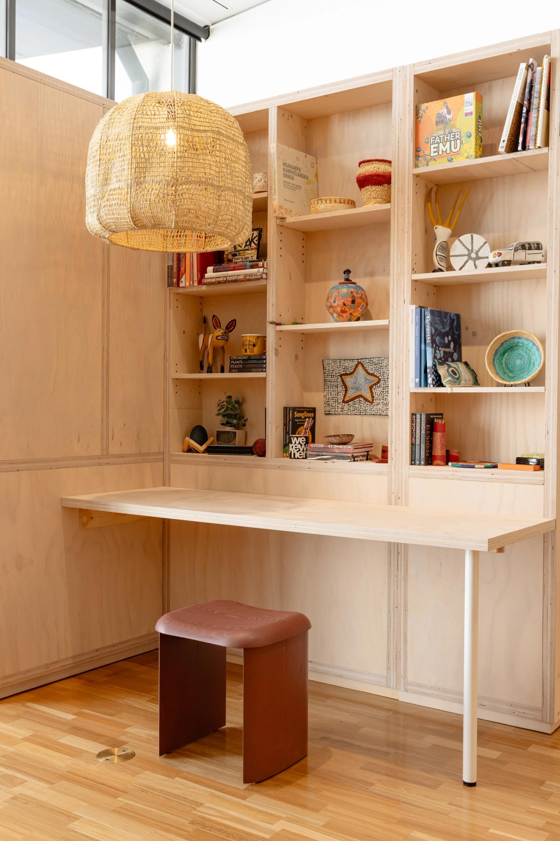 Study nook with light wooden desk, dark wooden stool, woven pendant light and shelving filed with items and books.