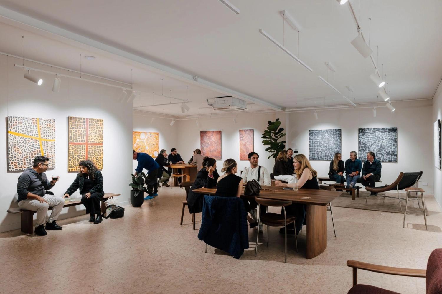 People sit in various groups on timber furniture in an art gallery