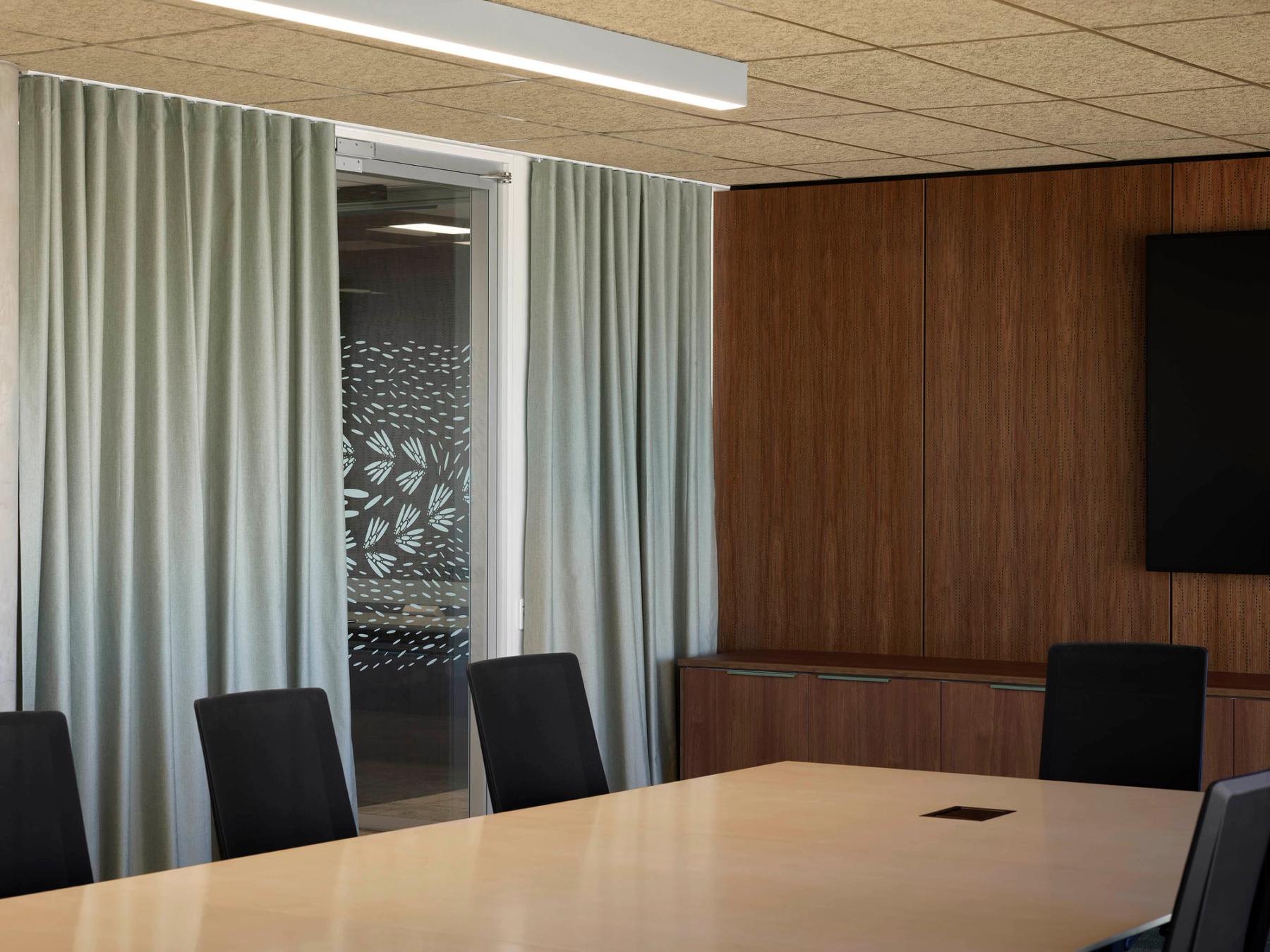 Meeting room with textured wooden panels, a long boardroom table surrounded by black chairs, curtains along the wall opened to show off decals.