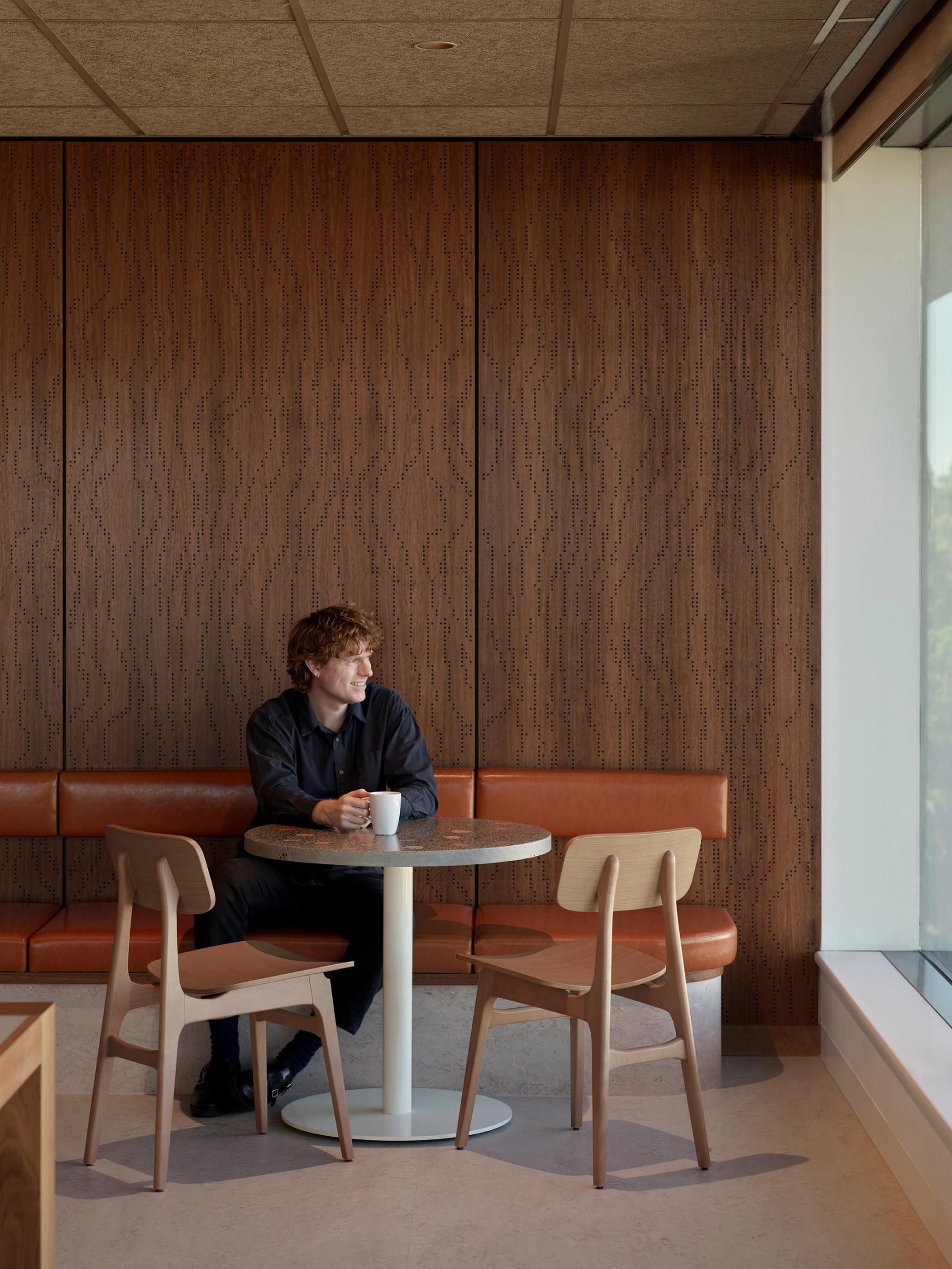 Man sitting at a small round table with a mug, wooden textured panel walls are behind him.