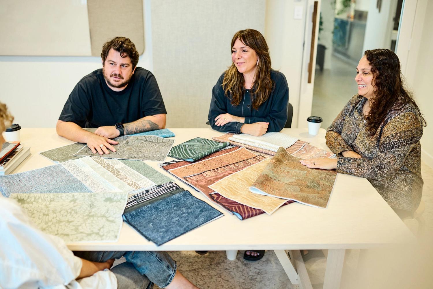 Co-owners Troy, Laetitia and Amanda sit at a table covered in textile samples looking towards co-owner Jessica