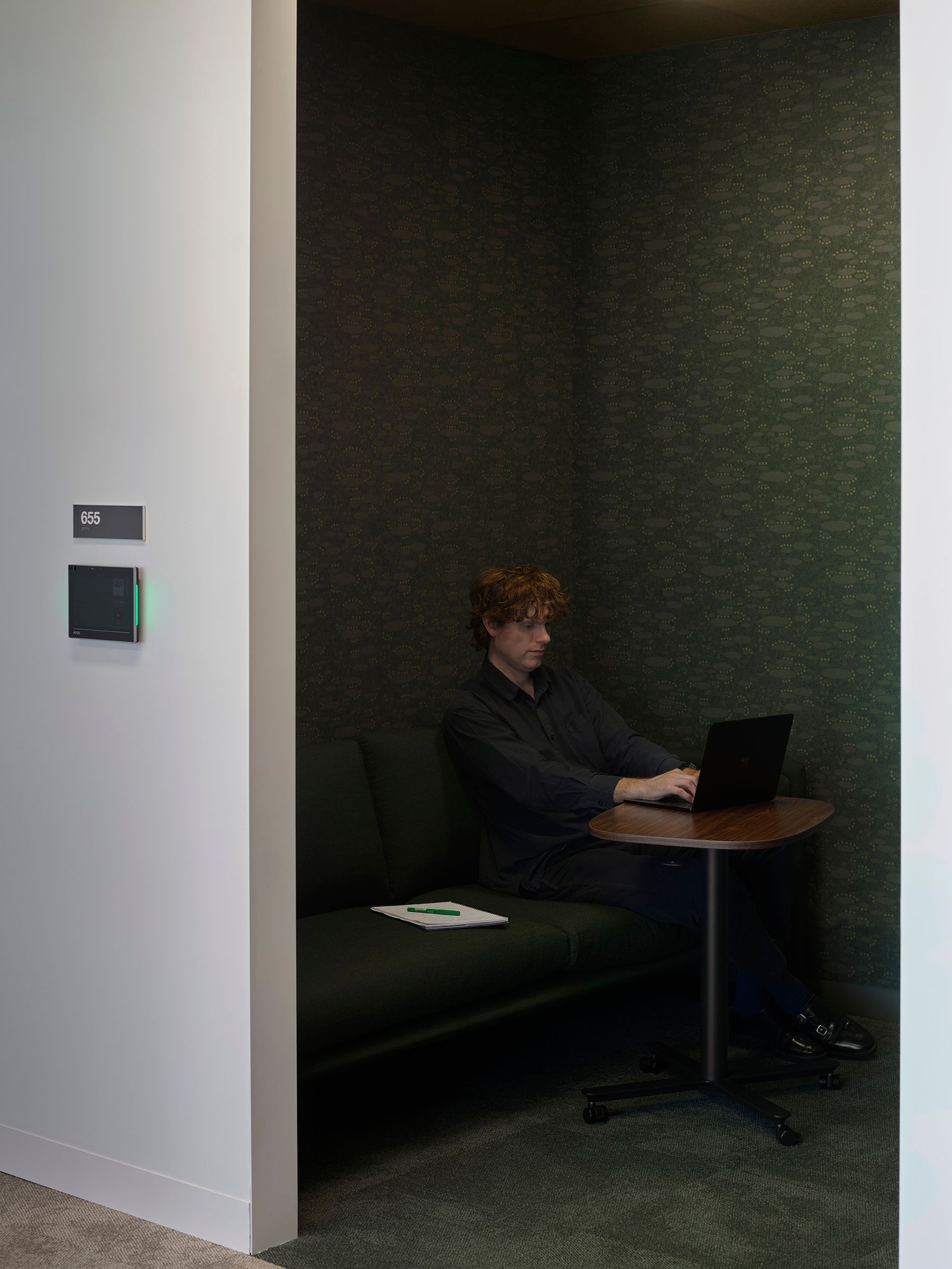 Man working on a laptop in a meeting room at a desk, there is textured green wall paper and green seating.