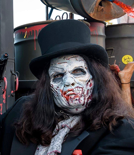 A zombie wearing a top hat with fake blood and wounds, standing near barrels with biohazard signs.