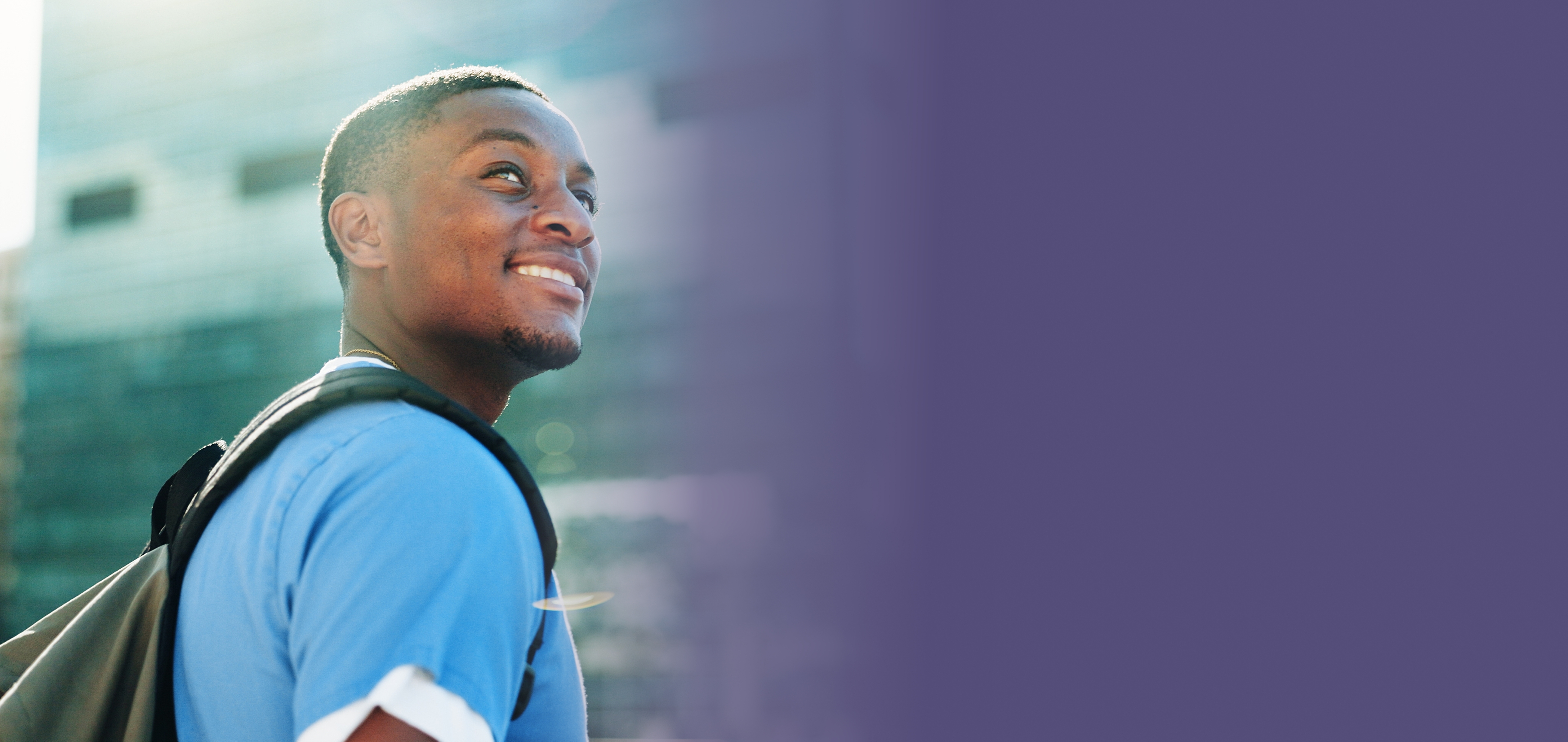Smiling Black man with backpack looking up; city building behind him; violet color field to the right.
