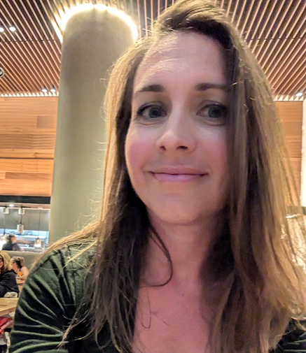 A woman smiles in a restaurant, with a large pillar and wood paneled walls and ceiling behind her.