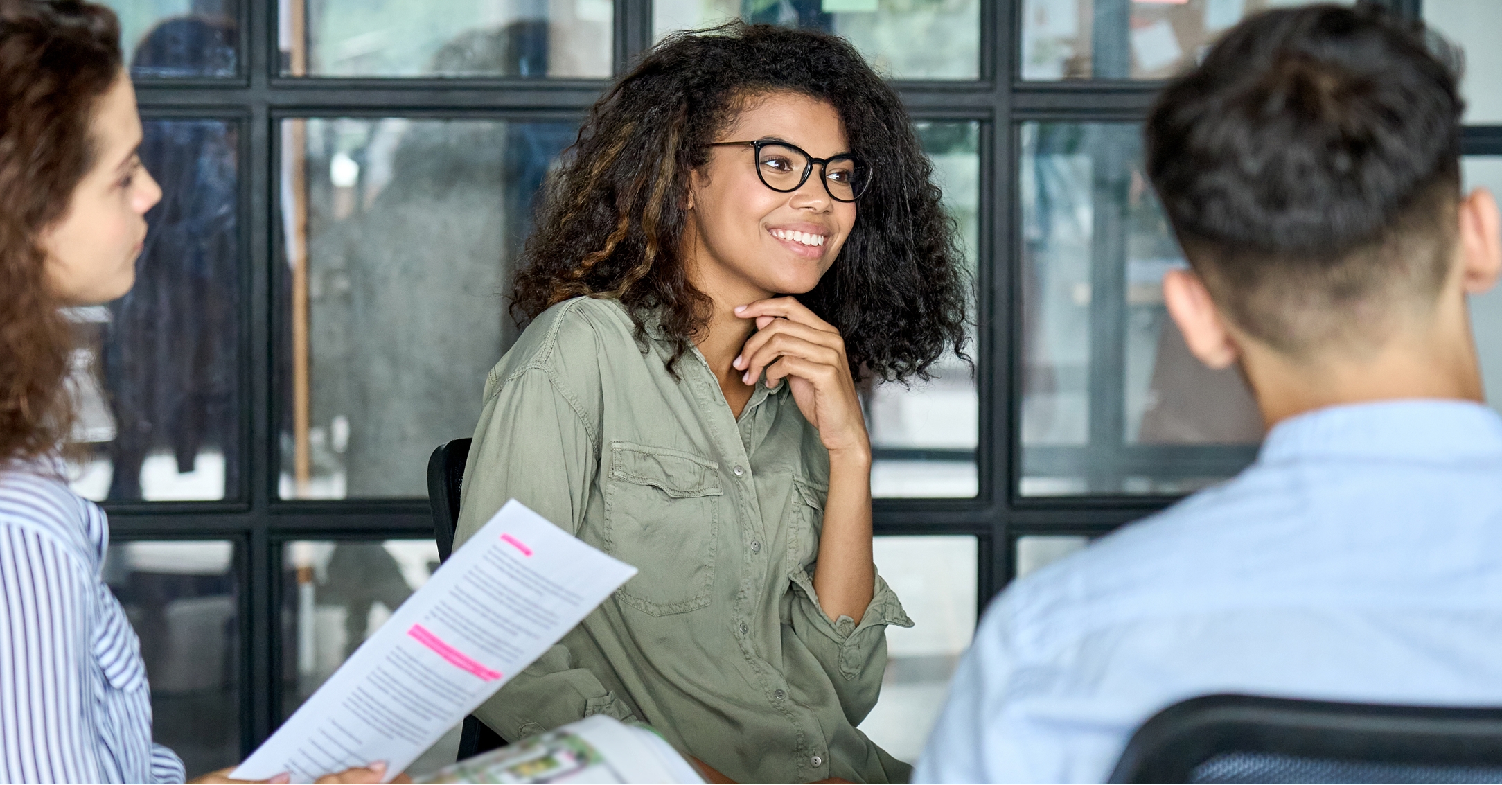 Three people sit in a meeting, with a smiling Black woman in glasses at the center. Documents are visible.