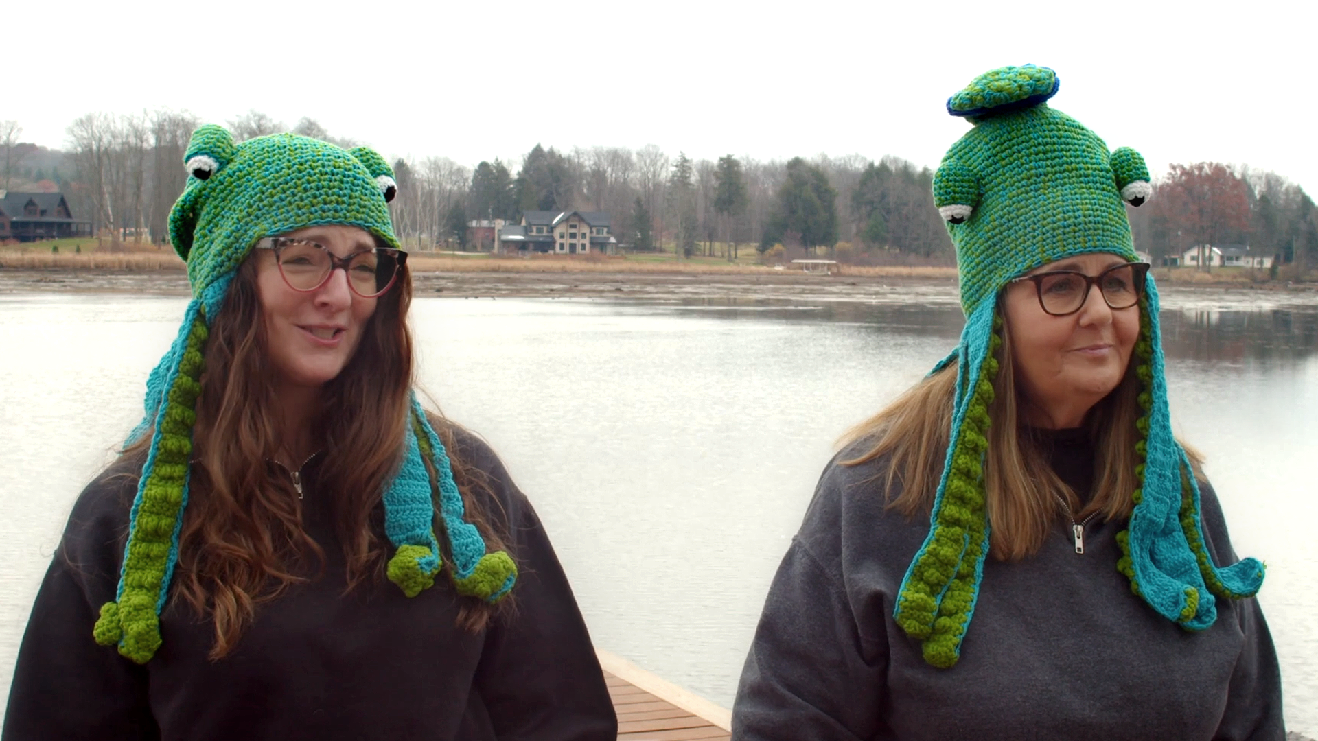 Two women wearing teal and green octopus hats stand on a dock by a lake.