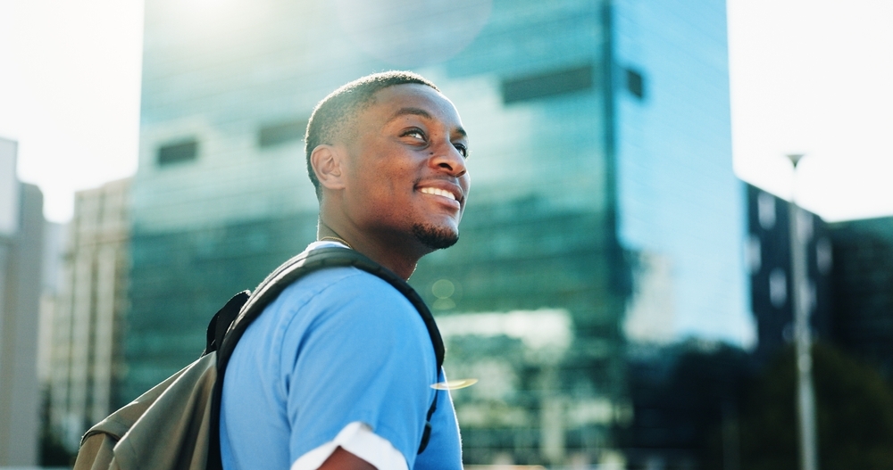 Image Smiling Black man with backpack looks up in city. Modern architecture in background.