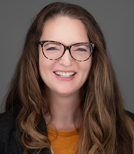 A smiling woman with long brown hair, wearing patterned glasses, a yellow shirt and a black blazer.