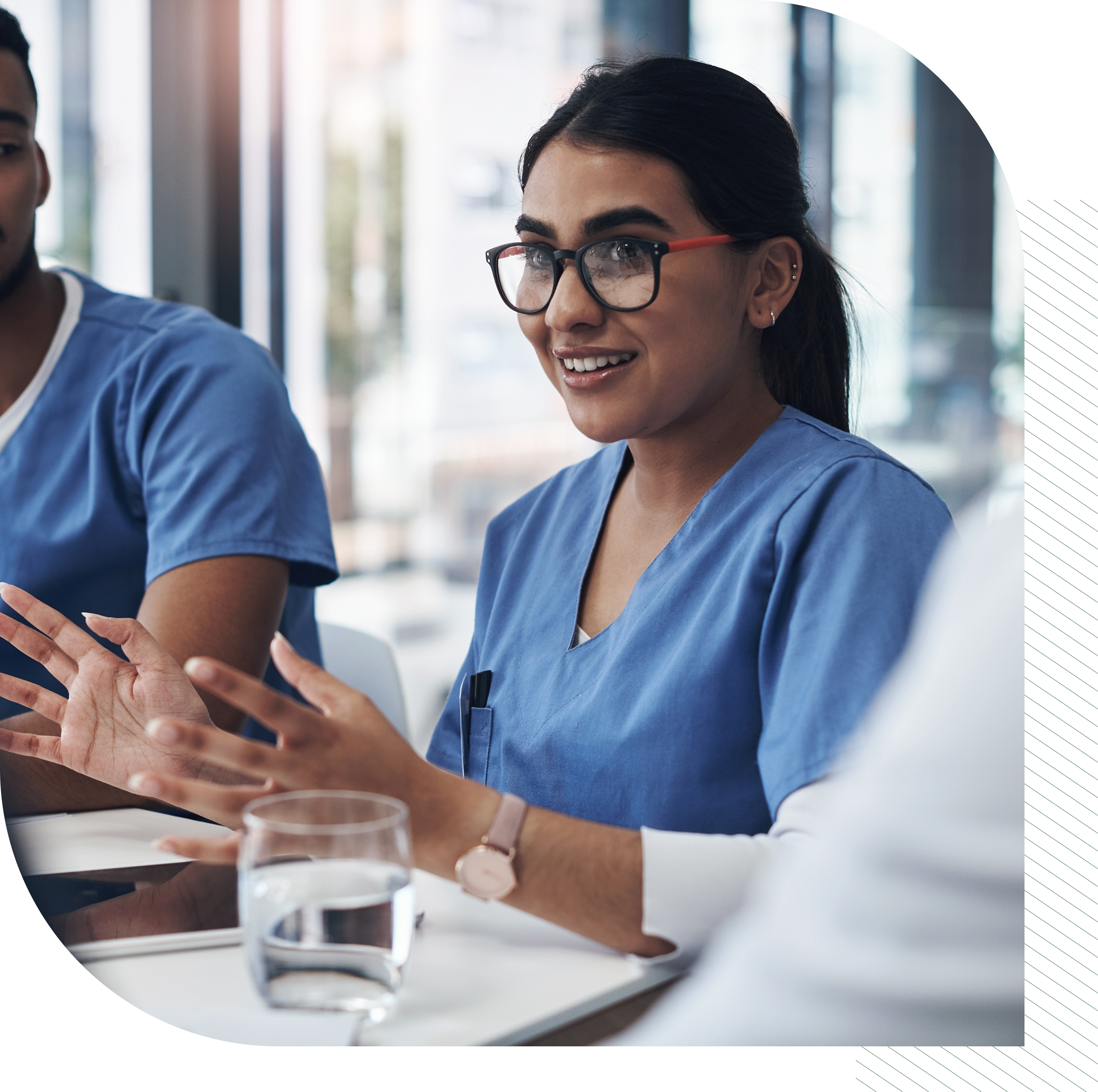 Medical staff in scrubs sit at a table, gesturing and talking during a meeting. A glass of water is on the table.