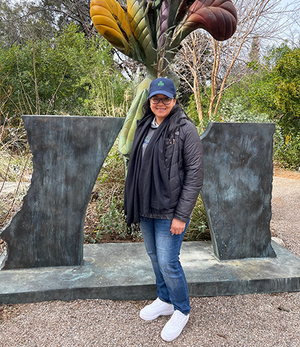 A woman in a jacket, jeans, and hat stands in front of an abstract sculpture in a garden.