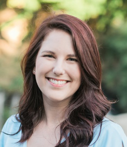 Smiling woman with auburn hair and light blue top, outdoors with blurred greenery in background.