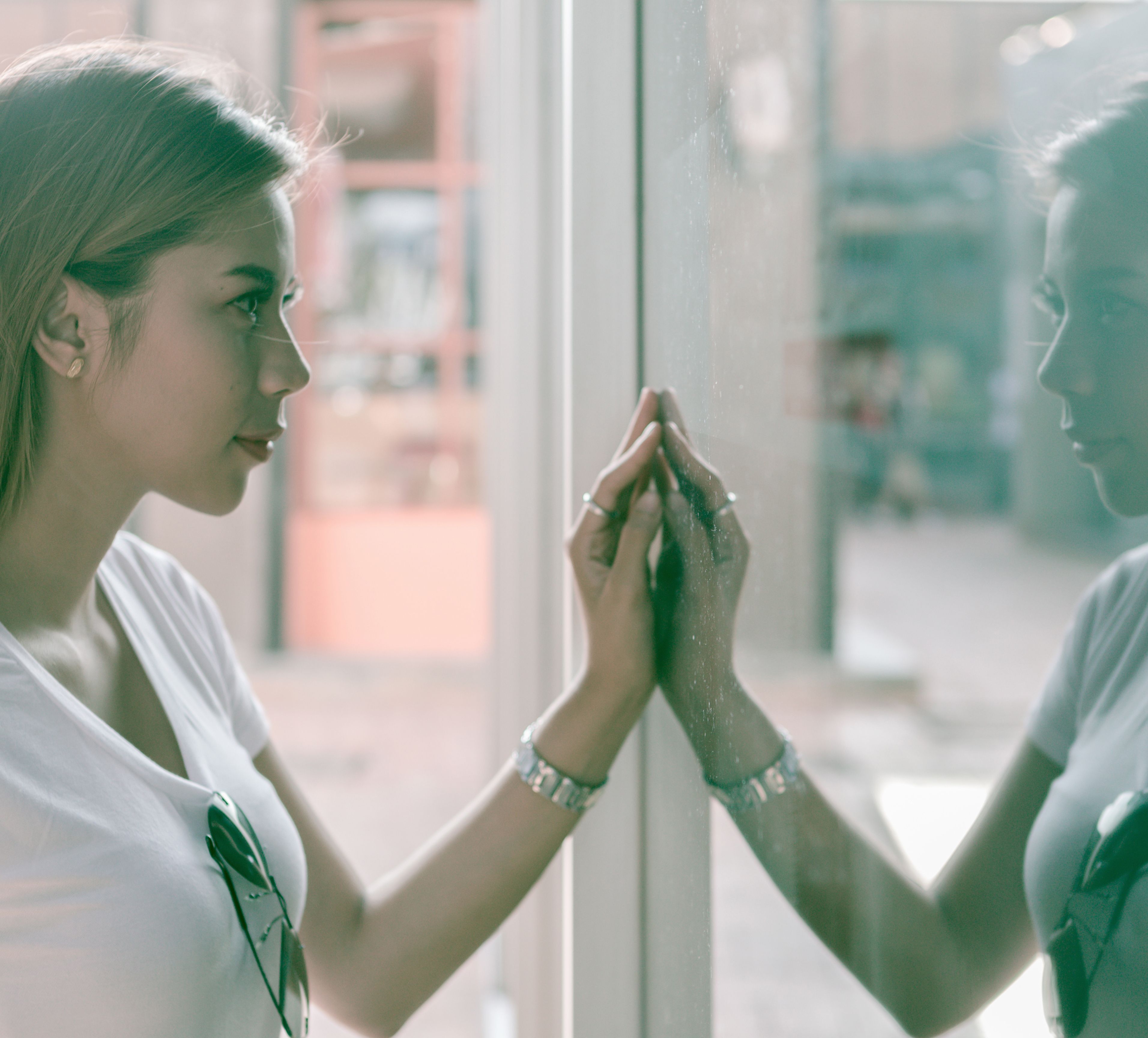 Woman with hand on glass looks at reflection, wearing a white t-shirt and bracelet.