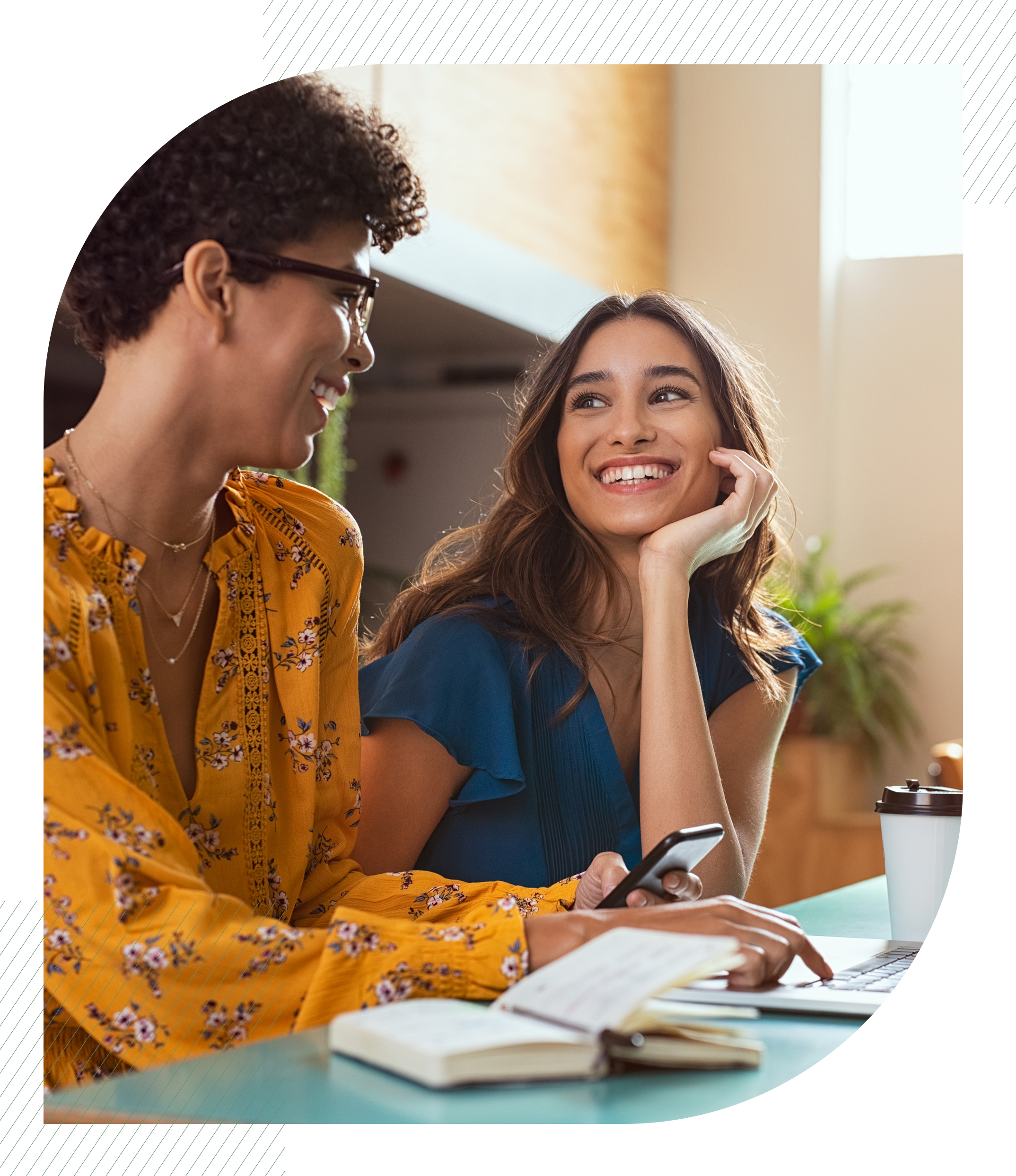 Two smiling women at a table with a laptop, phone, and notebook, appearing to collaborate.