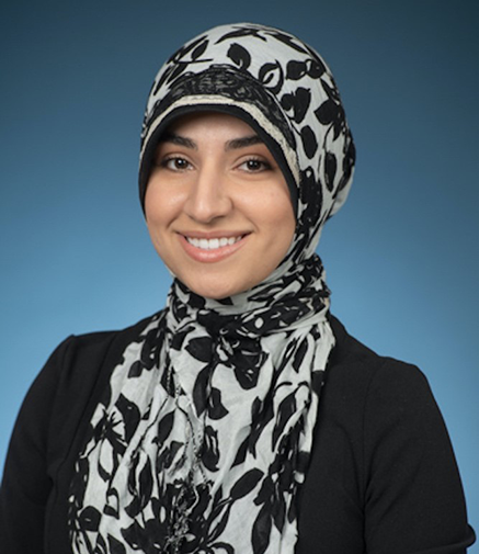Woman with hijab smiling, wearing black and white floral scarf against a blue background.