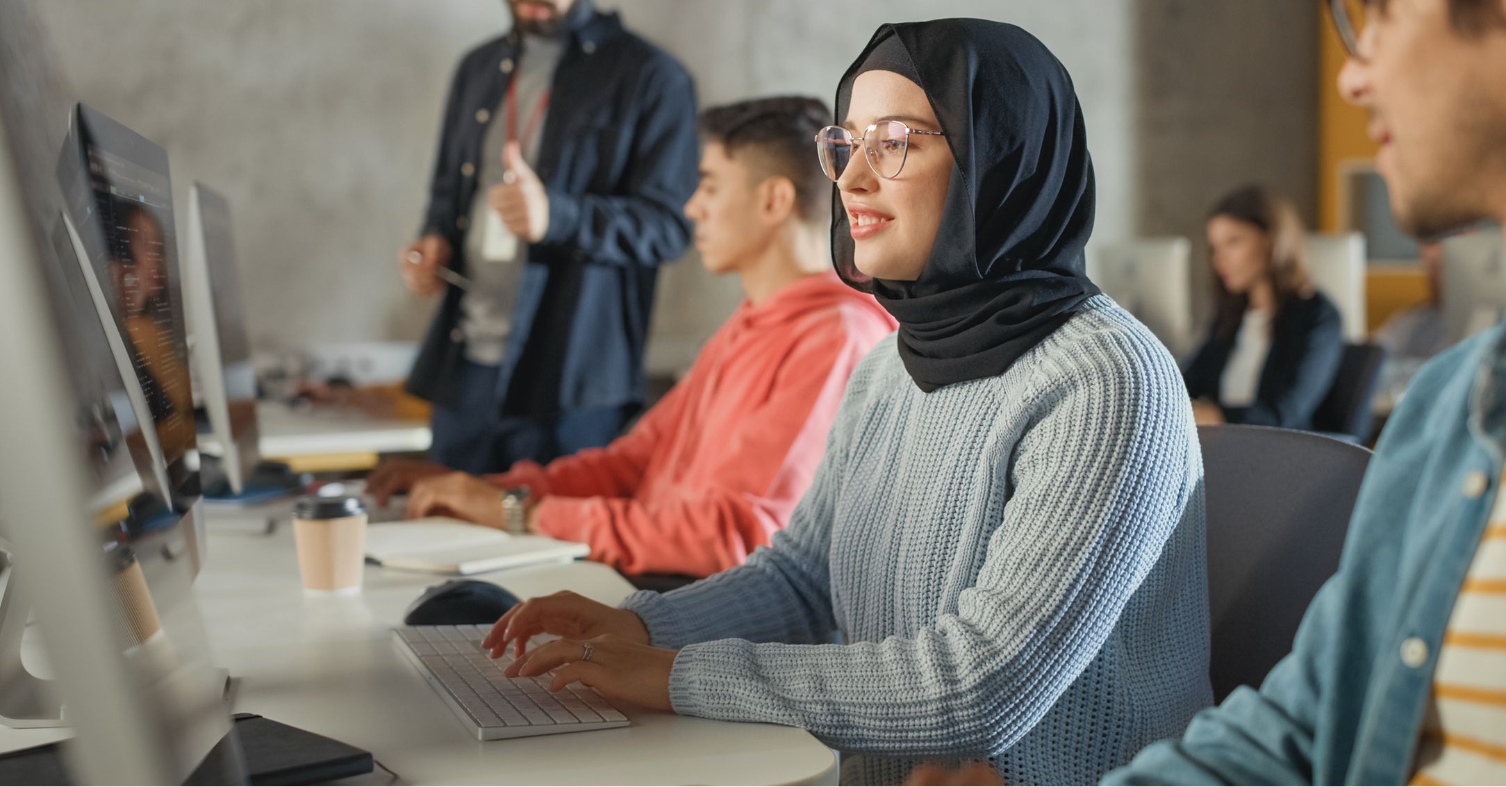 A young woman in hijab types at a computer in a classroom with other students and an instructor.