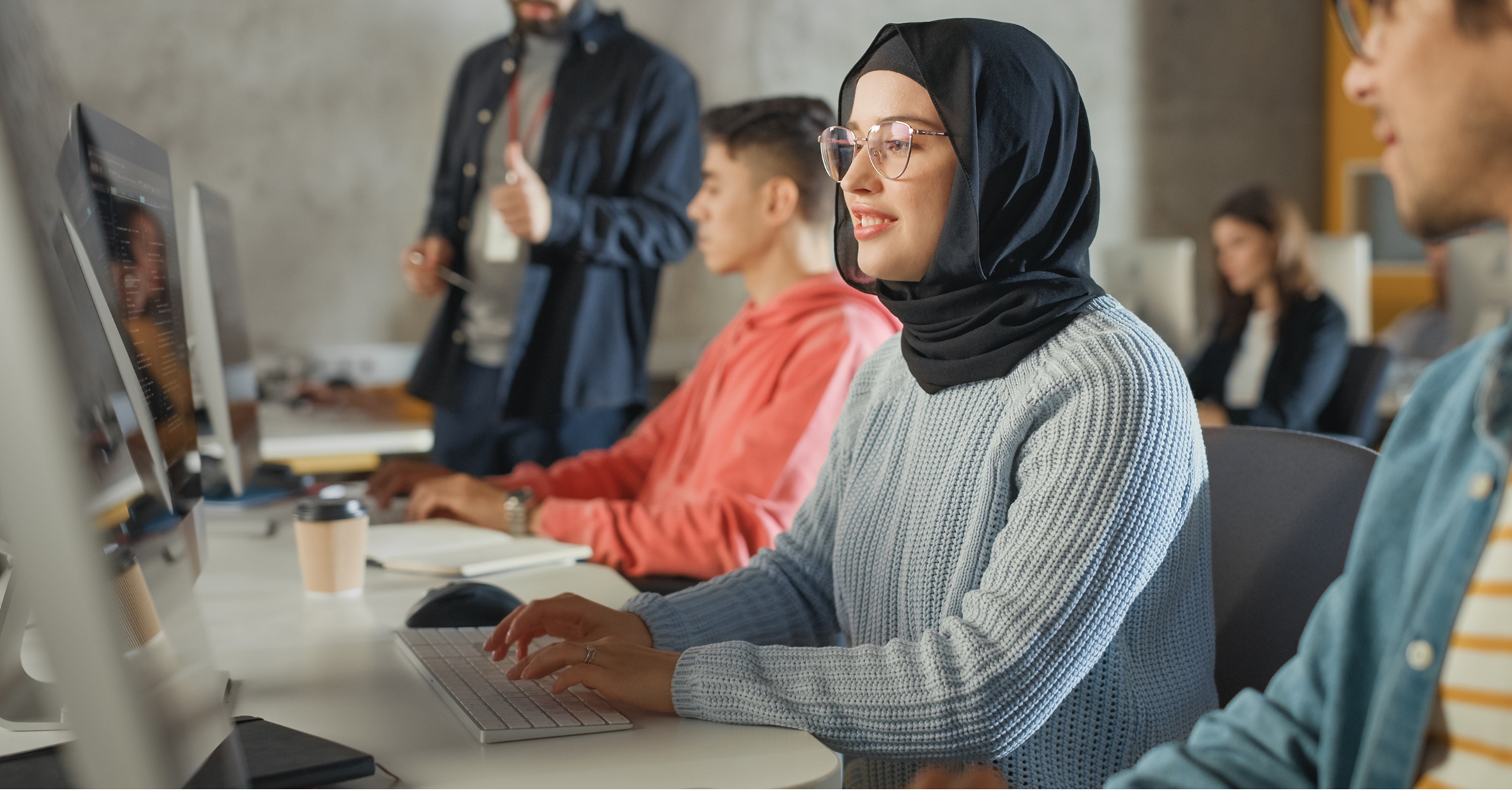 A young woman in hijab types at a computer in a classroom with other students and an instructor.