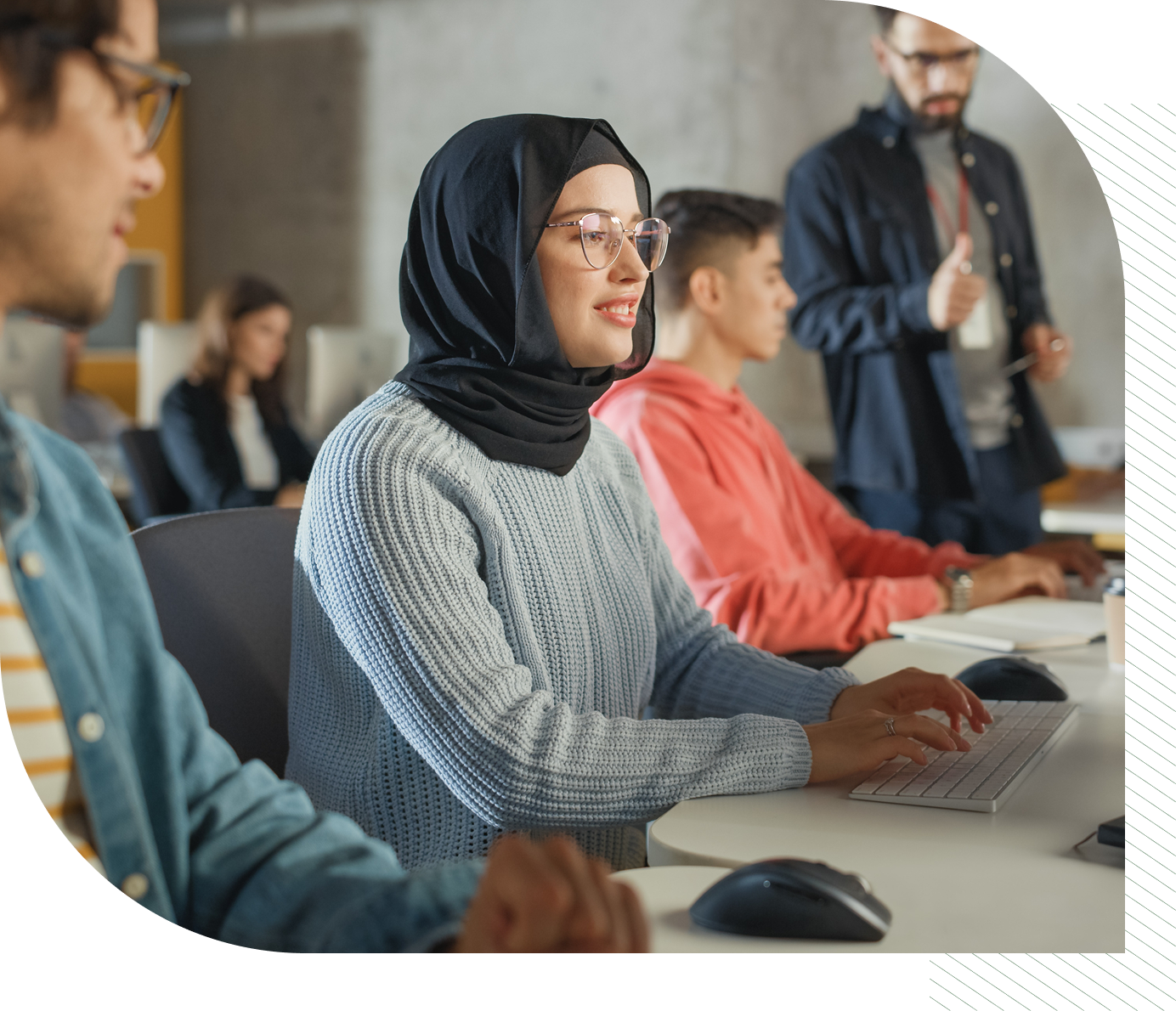 Students in a classroom setting, woman in hijab at computer, teacher in background.