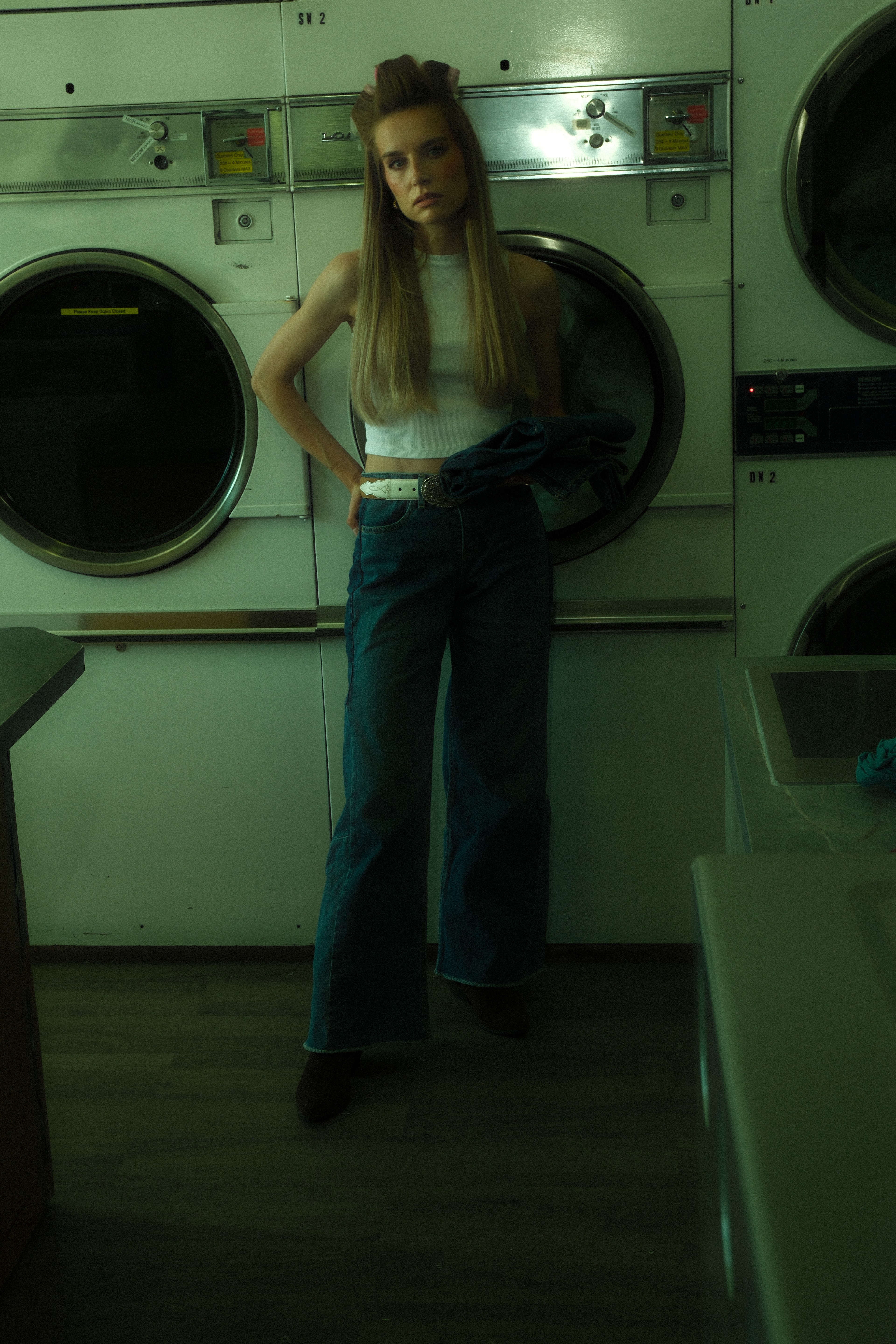 Woman wearing a white sleeveless top and wide-leg denim jeans standing inside a green-lit laundromat in Calgary.