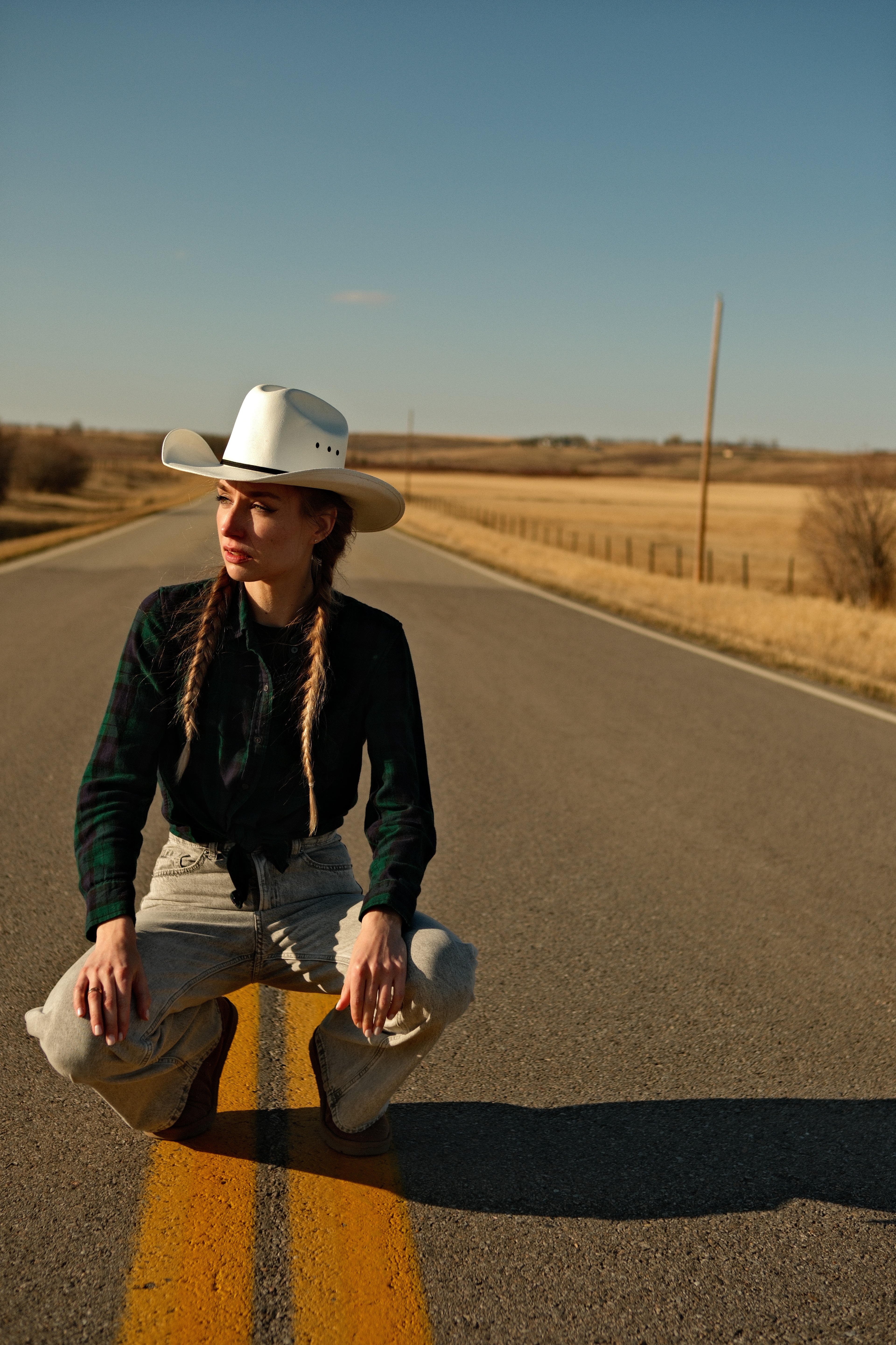 Model Mariana squatting on a rural highway, white cowboy hat and twin braids, sunset lighting.