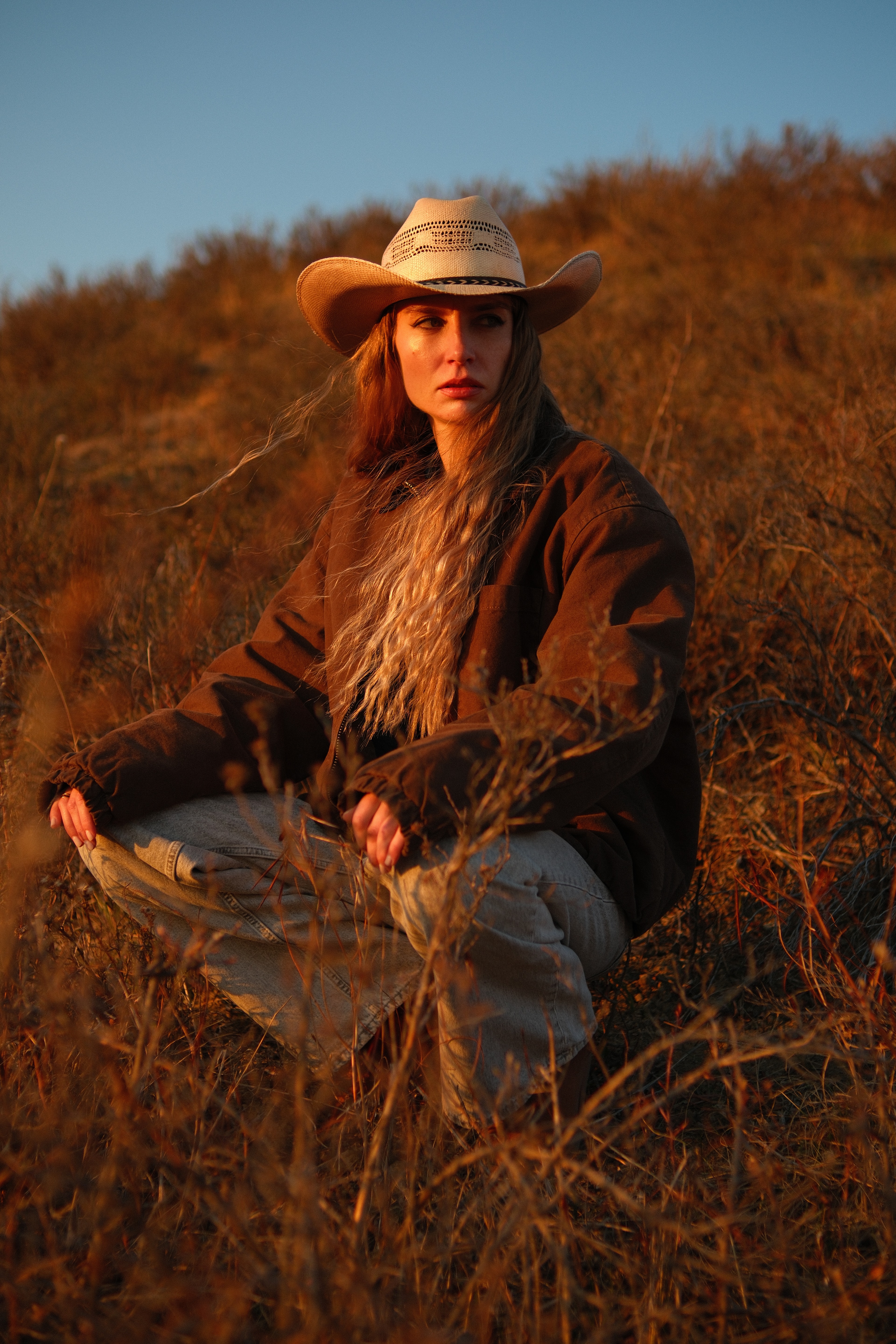 Golden hour portrait of model Mariana wearing a cowboy hat, seated in open fields at sunset.