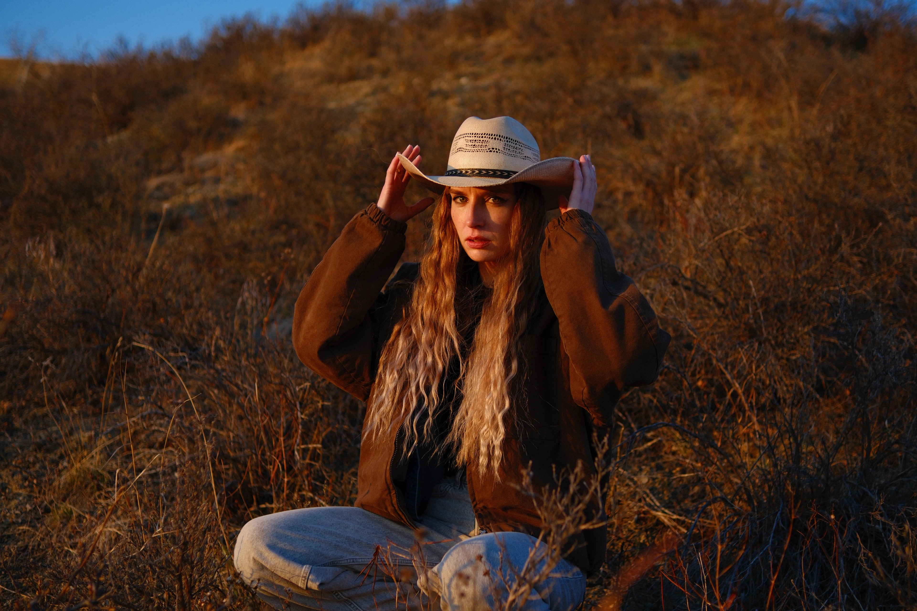 Rustic golden hour portrait of model Mariana adjusting her cowboy hat in open fields, sunset photography