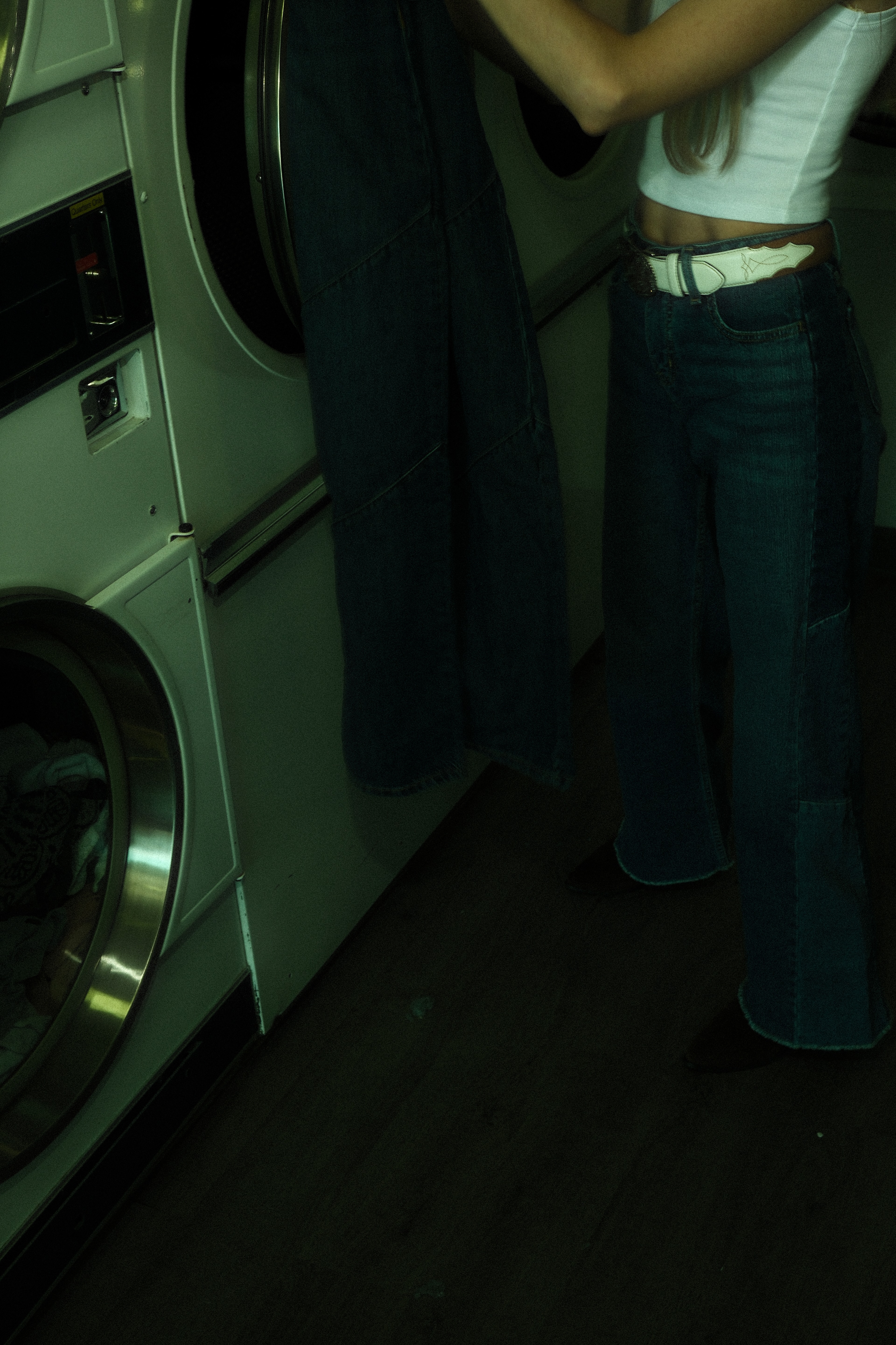 Woman in a white crop top and dark wide-leg denim leaning beside washing machines in a green-lit Calgary laundromat.