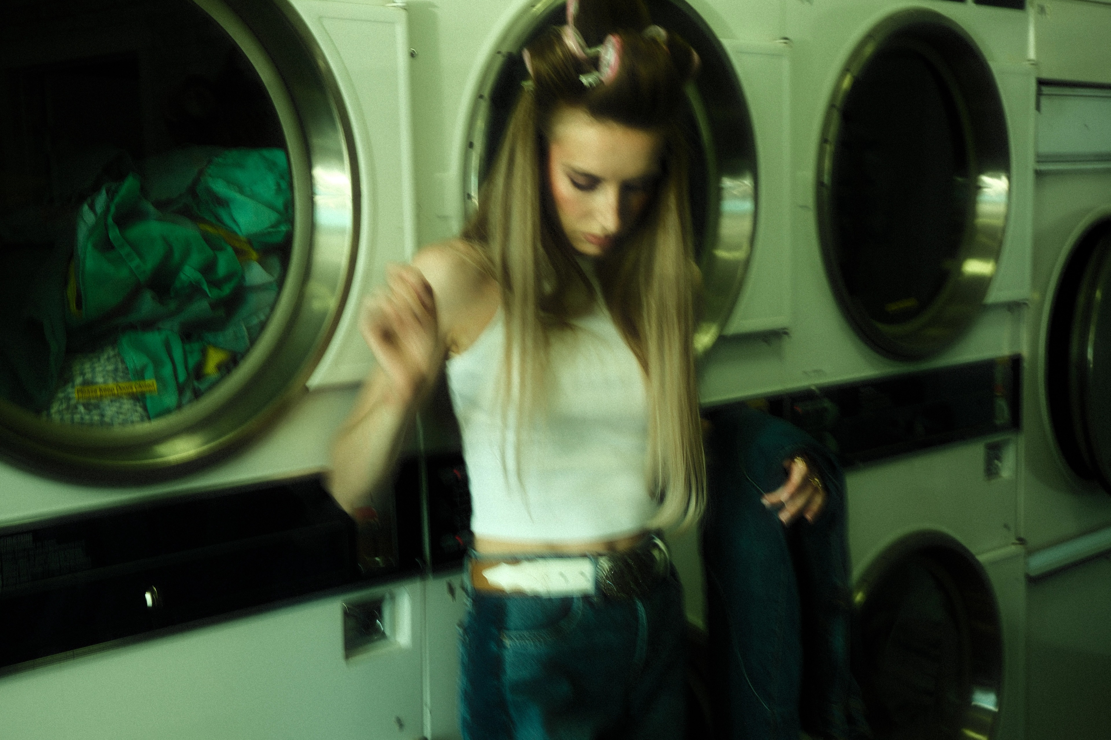 Woman wearing a white sleeveless top and wide-leg denim jeans standing inside a green-lit laundromat in Calgary.