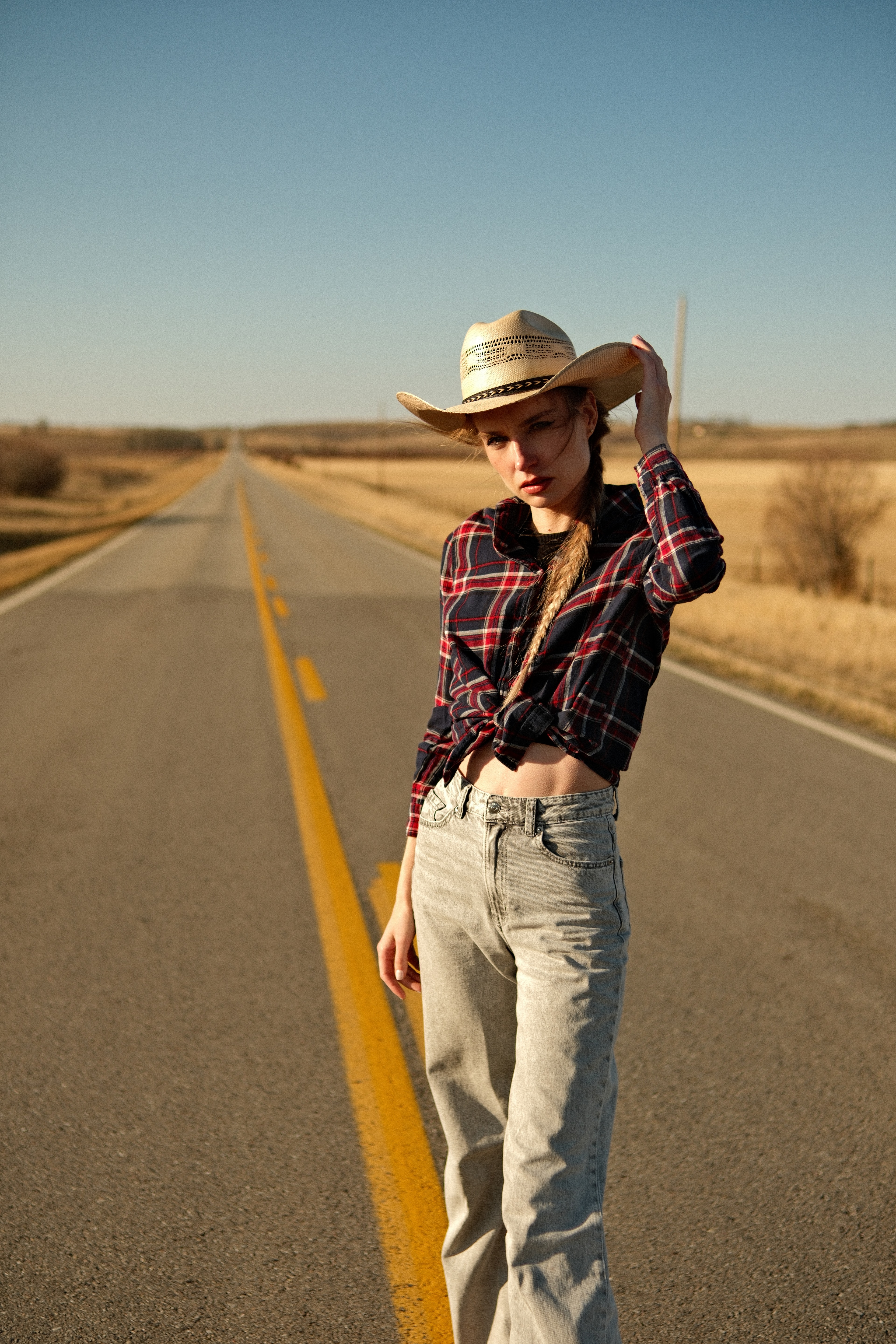 Outdoor portrait of model Mariana standing on an open highway, wearing cowboy hat and plaid shirt, natural sunlight.