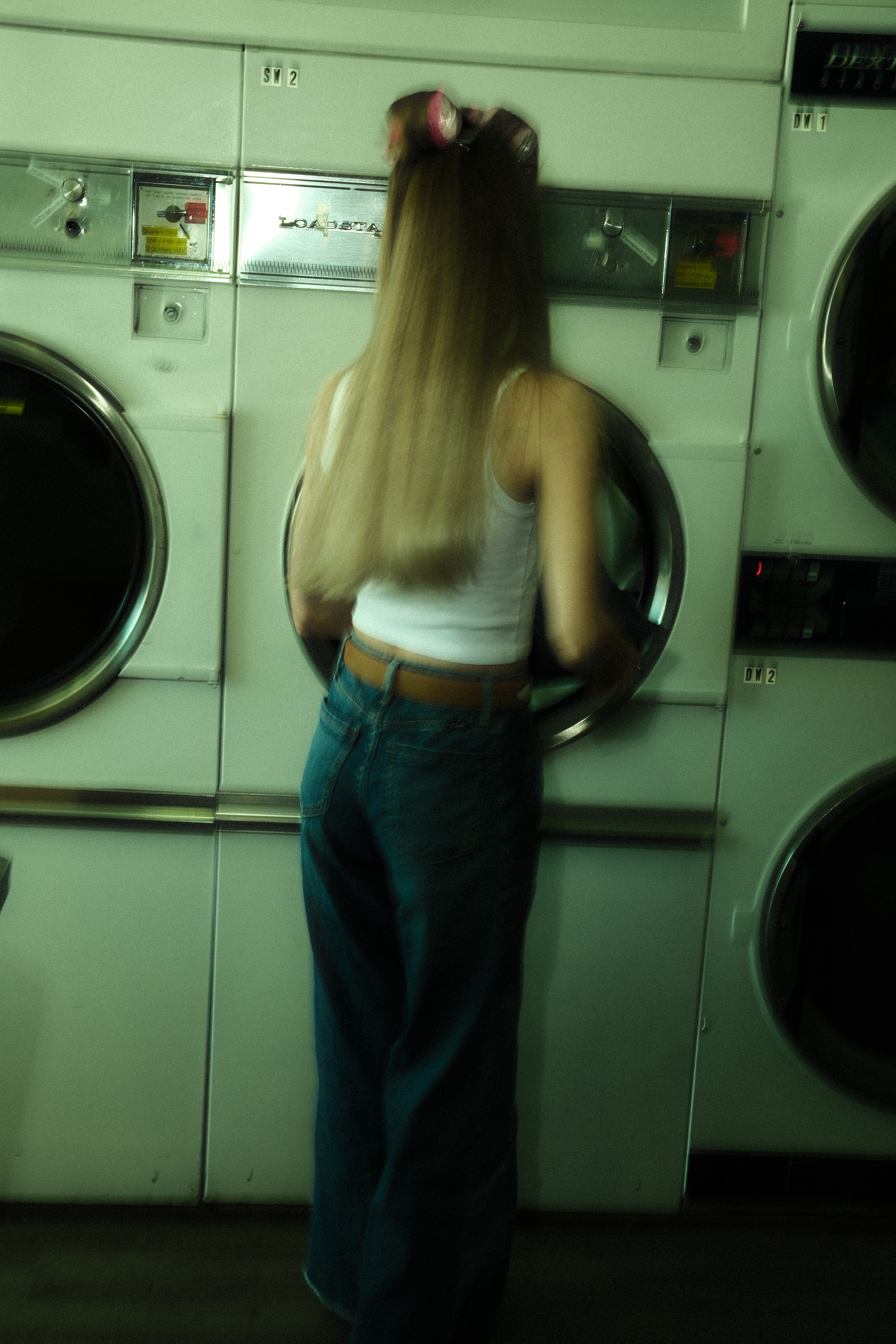 Woman wearing a white sleeveless top and wide-leg denim jeans standing inside a green-lit laundromat in Calgary.
