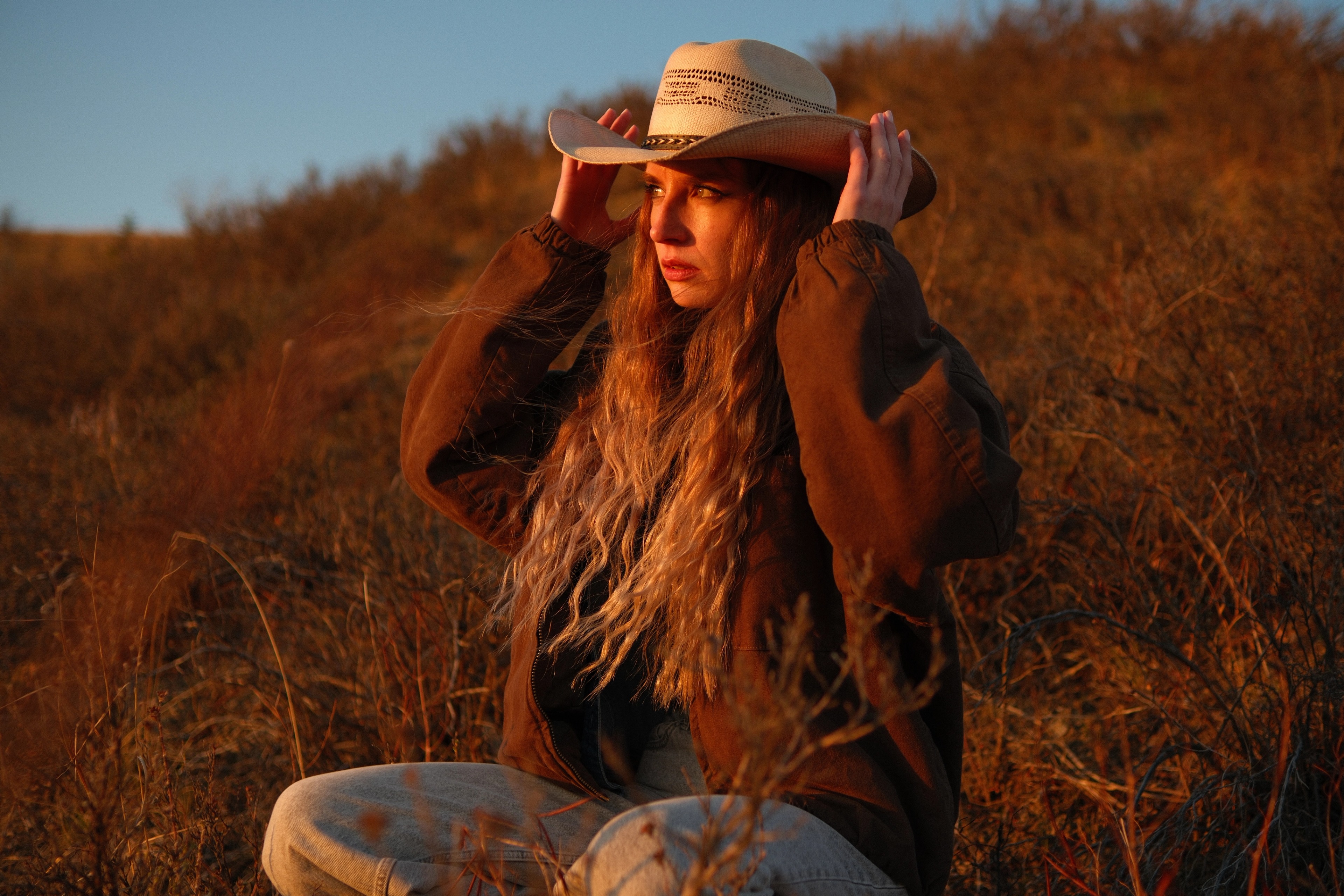 Golden hour side portrait of model Mariana in cowboy hat, surrounded by rustic dry fields at sunset.