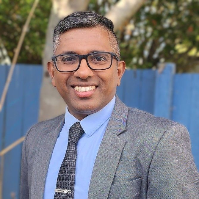 Saji standing with a grey suit and a blue shirt, black tie while smiling