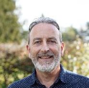 Man with gray short hair and lighter gray beard wearing a light teal and white t-shirt and smiling.