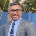 Saji standing with a grey suit and a blue shirt, black tie while smiling