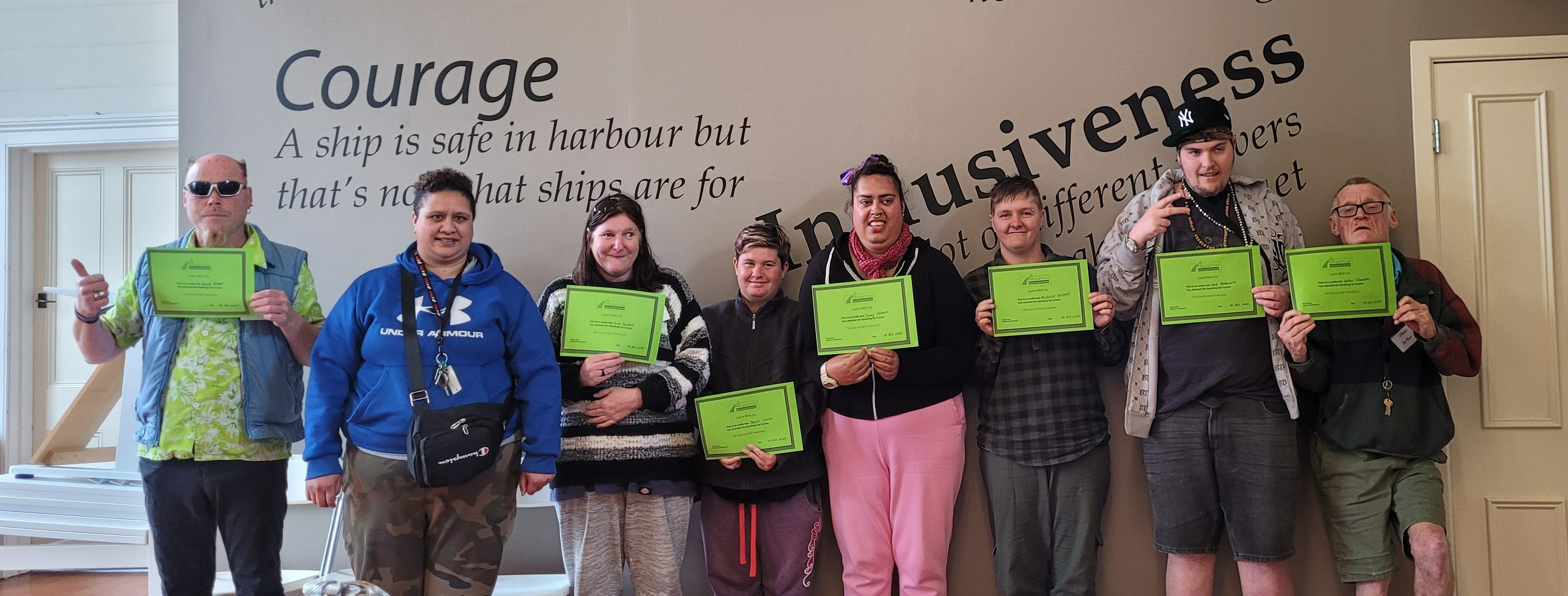 Line up of 8 people holding up green certificates in a room with a back drop that says Courage a ship is safe in harbour but that's not what ships are for.