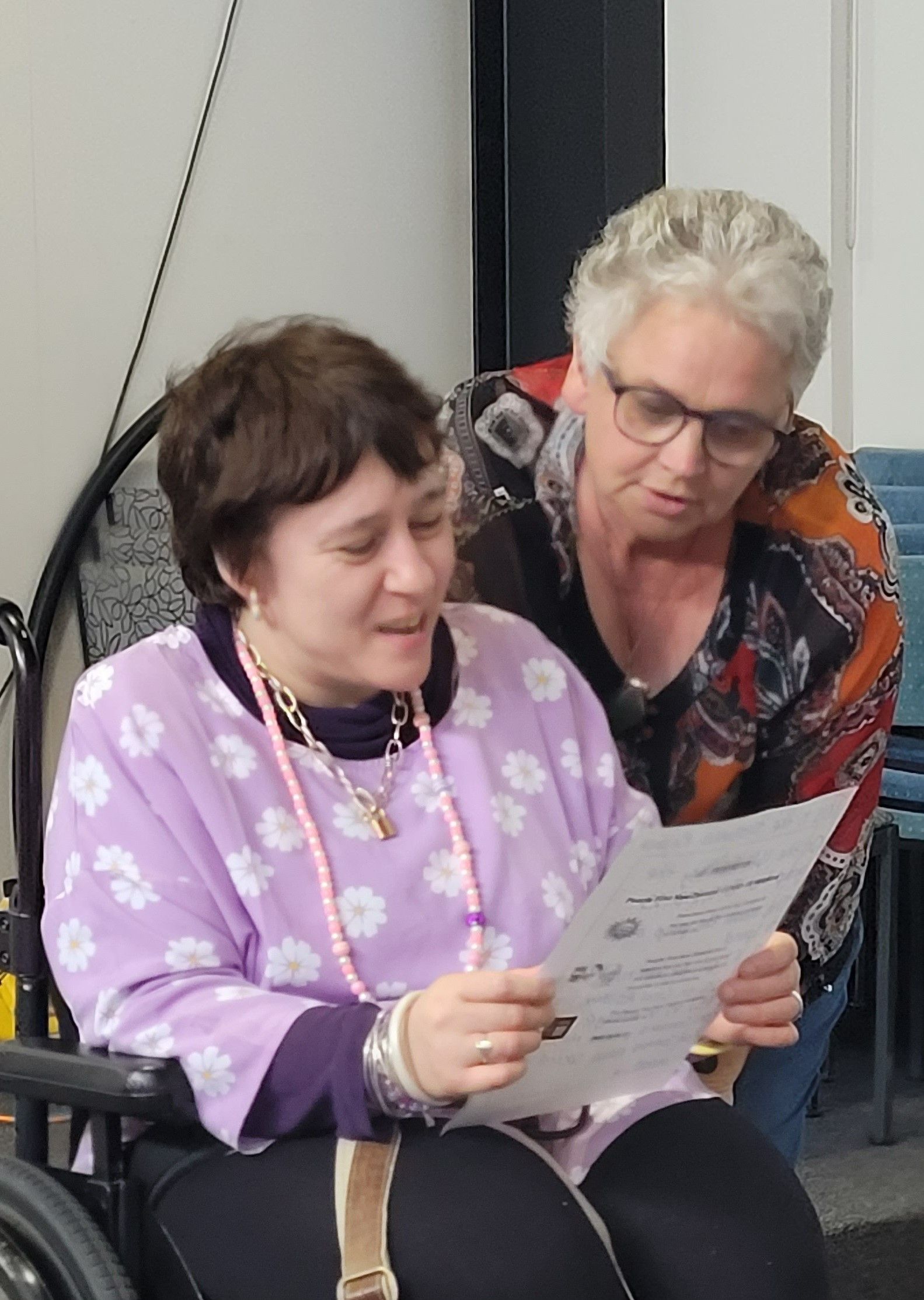 Two woman inside with the one on the left in a wheelchair wearing a pink top with white flowers, she has brown hair and is reading from a piece of paper, the second woman is on the right looking over the other woman's shoulder, she has grey short hair and glasses and is wearing an orange and black top.