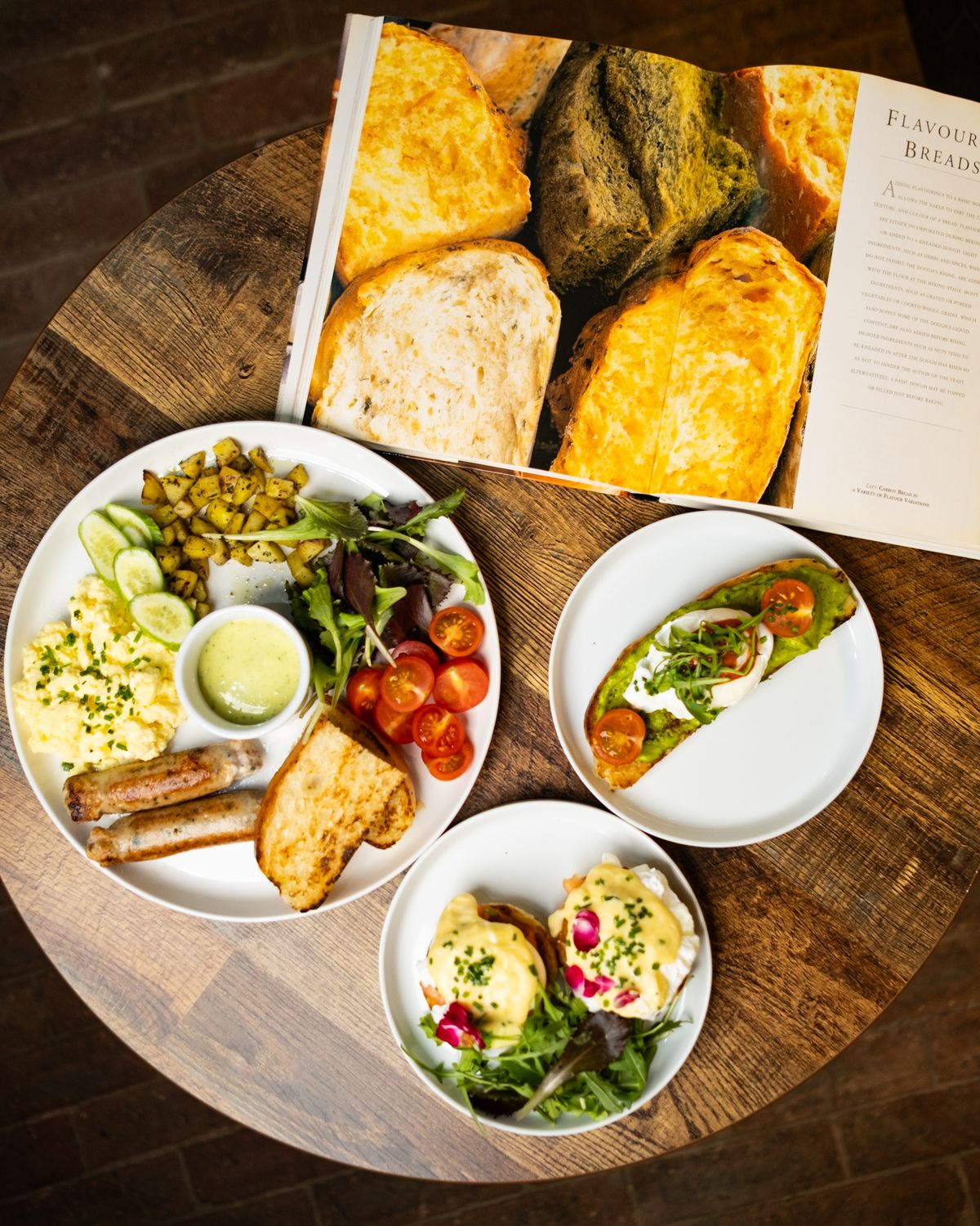 An overhead view of a full Lantees breakfast spread on a wooden table, featuring scrambled eggs, sausages, avocado toast, eggs benedict, and a fresh salad alongside an open bread cookbook