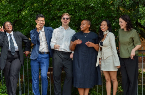 A diverse group of six people laughing and smiling outdoors against a green leafy background.