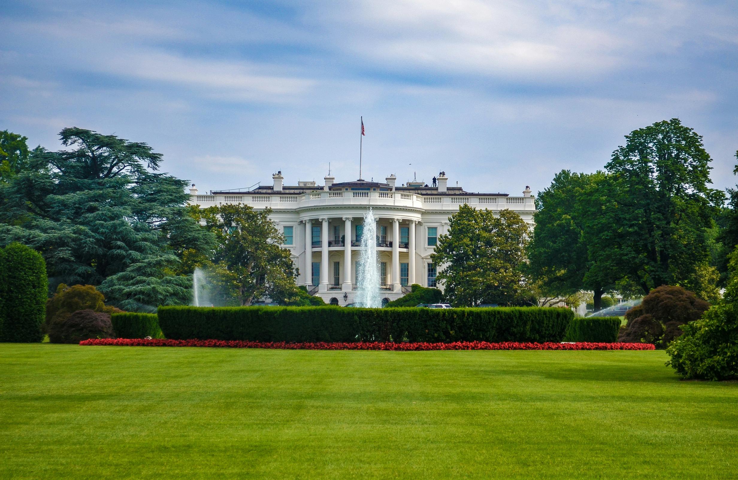 the white house is surrounded by trees and bushes and has a fountain in front of it .