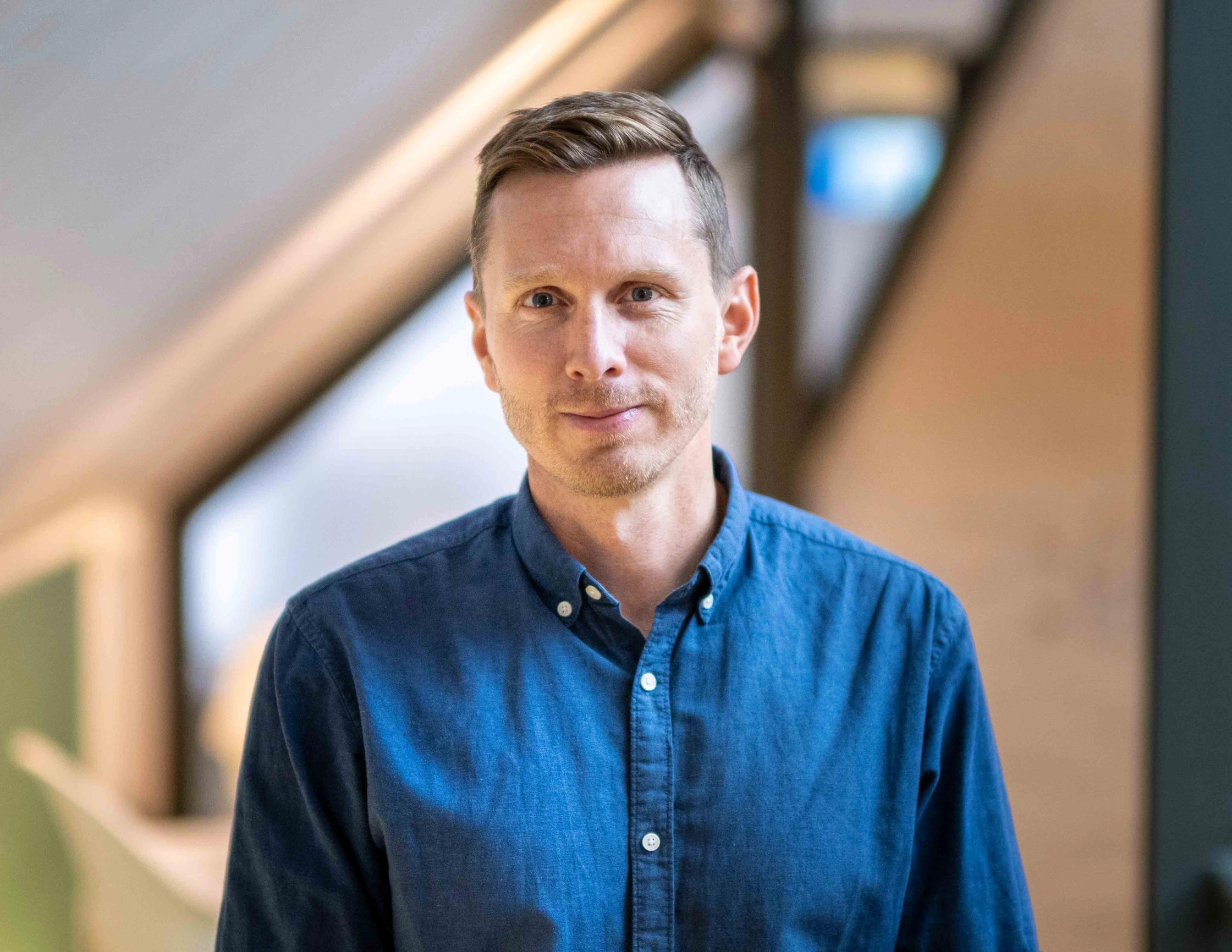 A man with short brown hair wearing a blue shirt looks directly at the camera.