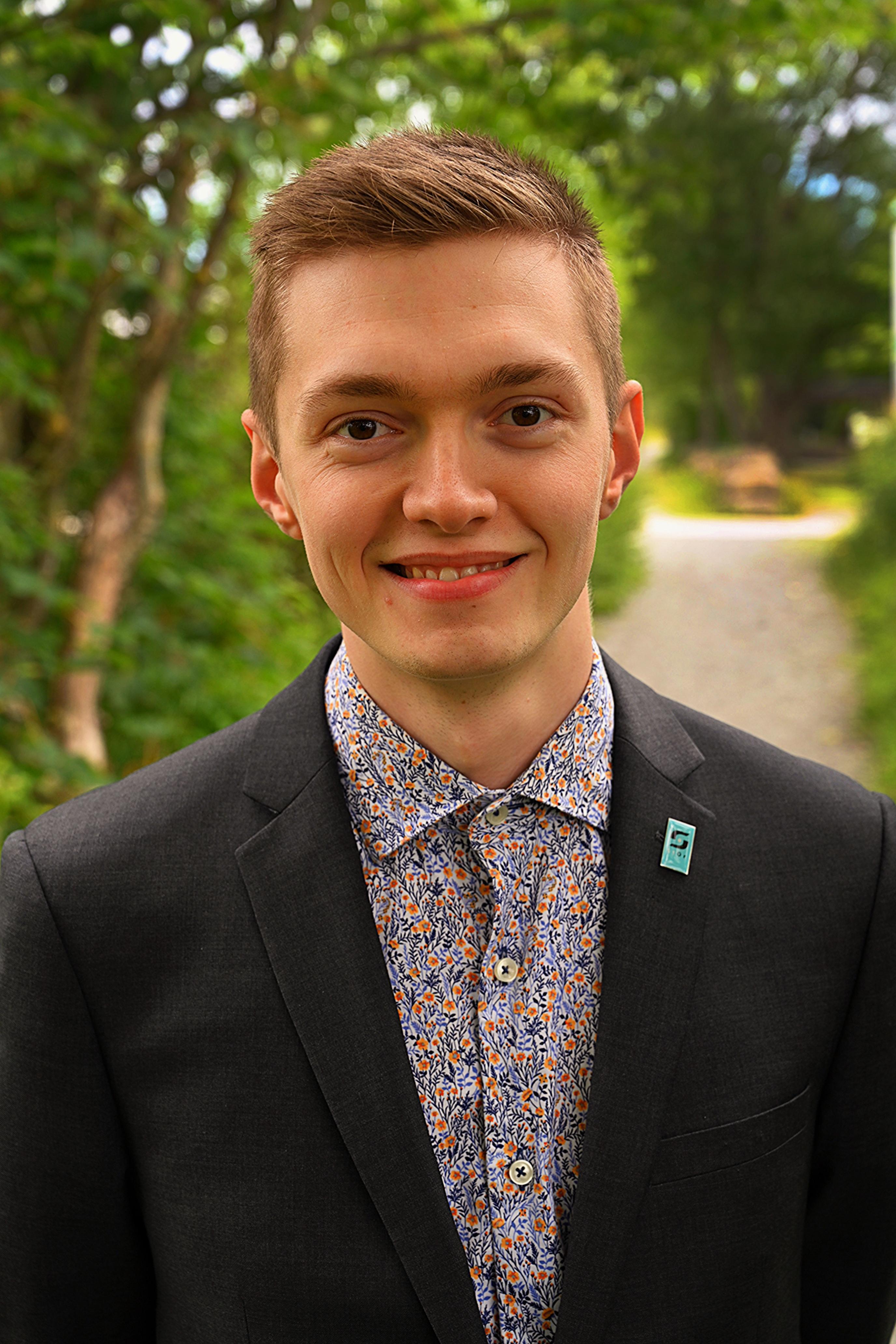 A smiling young man in a dark suit and patterned shirt, with a lapel pin, stands outdoors among green foliage.