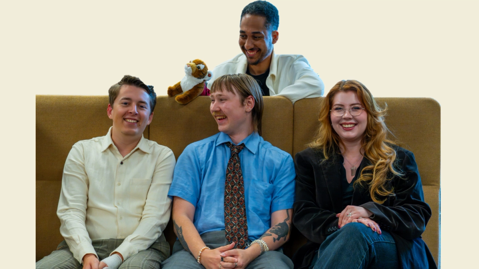 Four smiling people, three seated on a couch and one leaning over the back, with a small stuffed animal.
