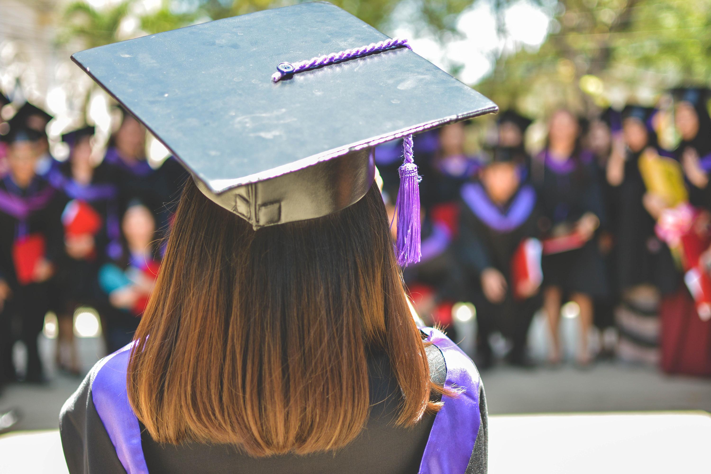 A graduate holds her diploma while looking out over a crowd.