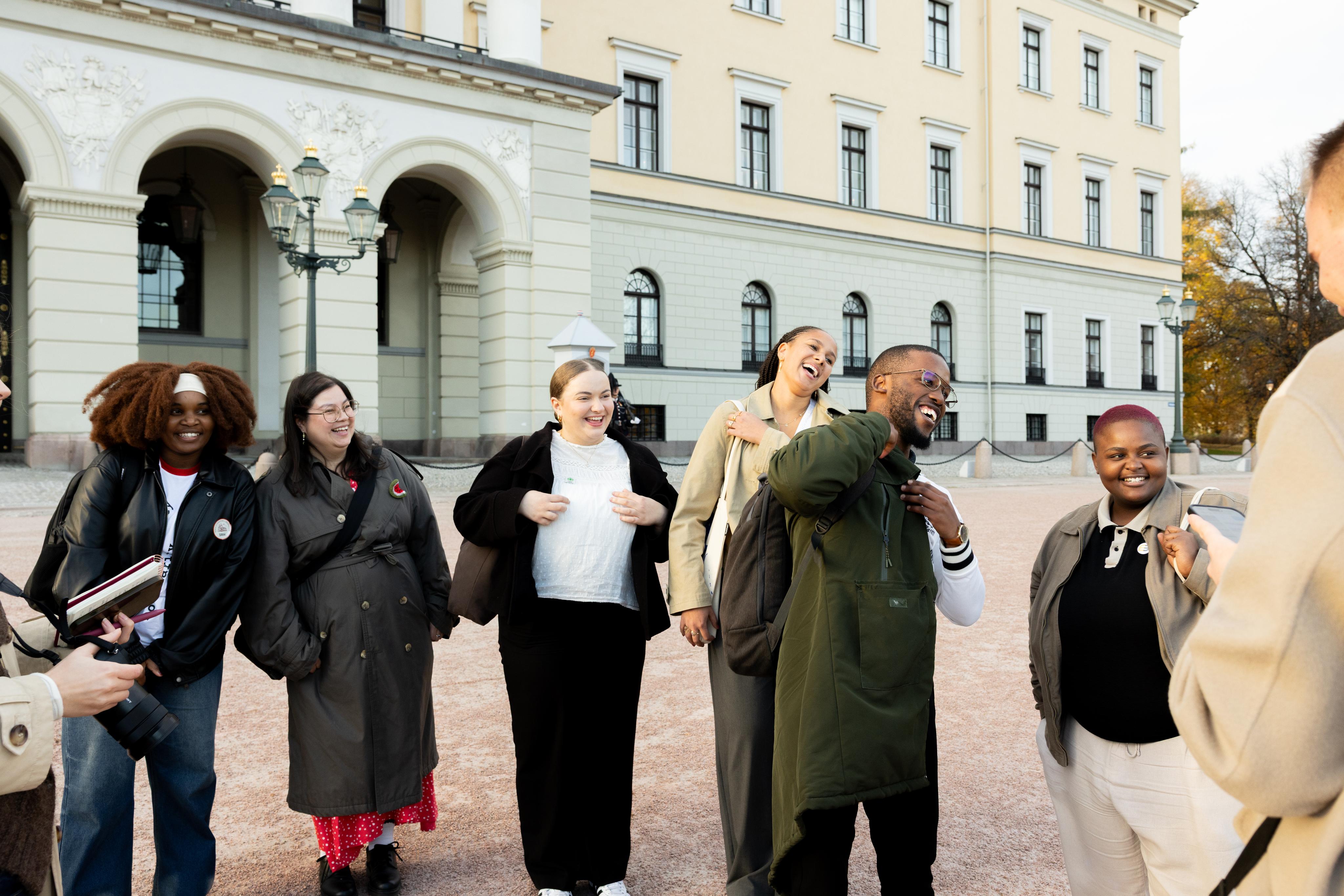 A diverse group of six young adults laugh and smile outdoors in front of a large, ornate building.