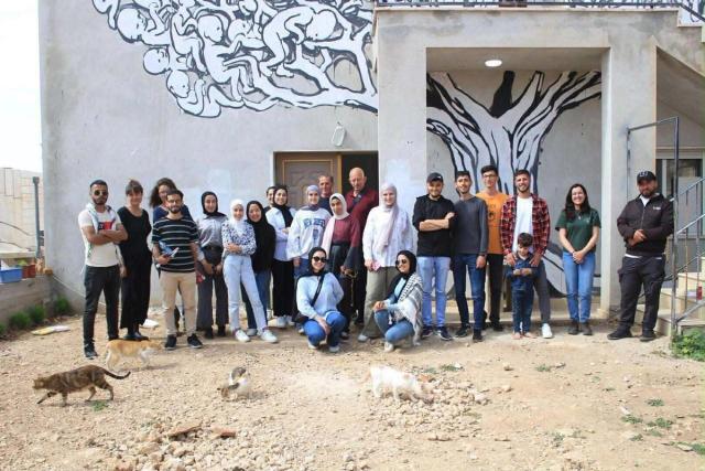 A diverse group of people and a child pose outdoors in front of a building featuring large murals of a tree and abstract figures, with several cats on the dirt ground.