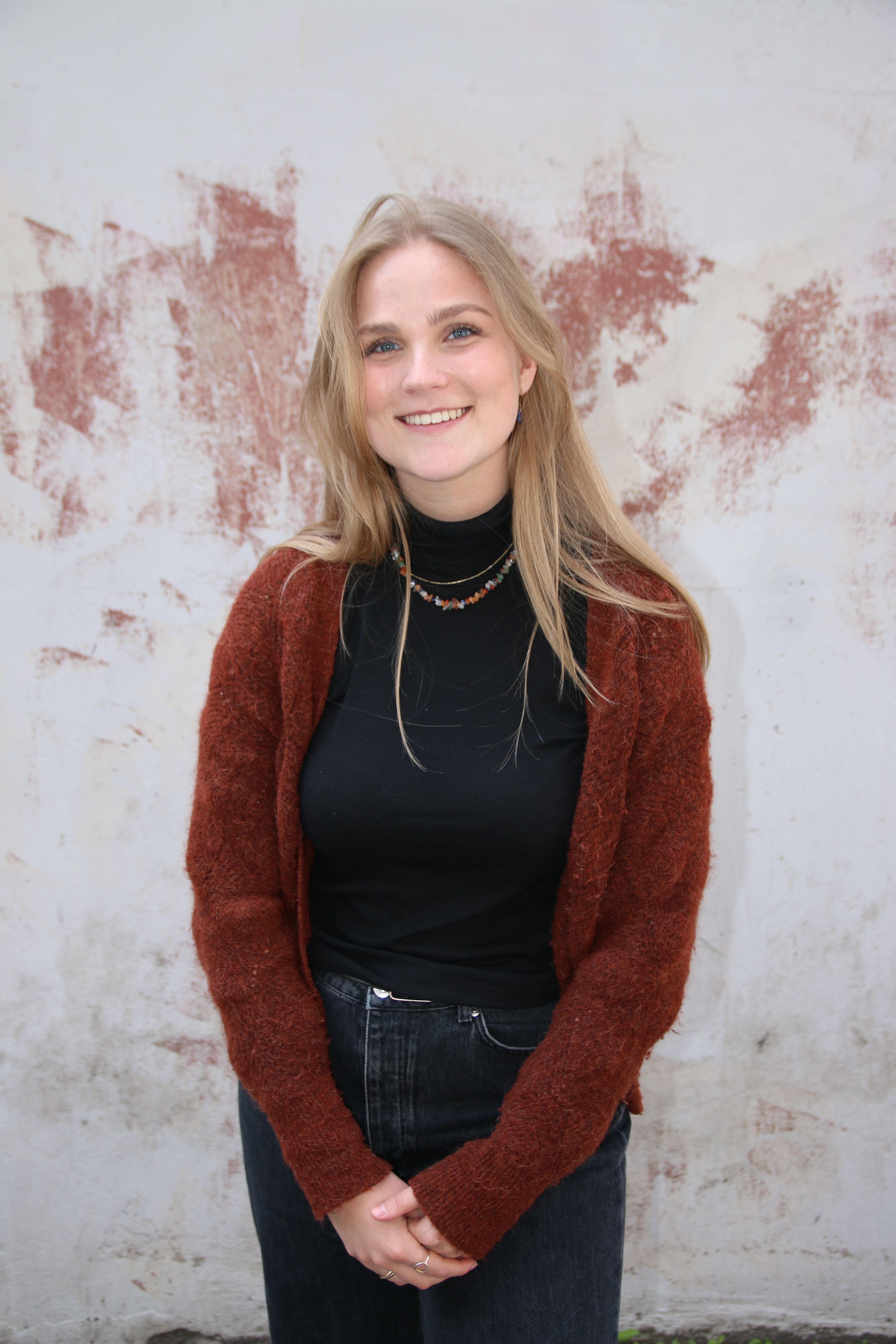 A smiling young woman with blonde hair wears a black turtleneck and a rust-colored cardigan against a textured wall.